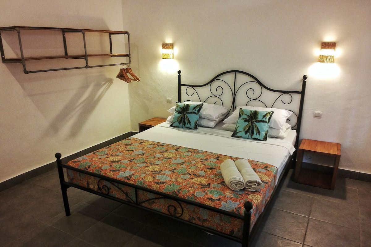 Uma cama bem arrumada com um cobre-leito floral, duas almofadas e uma toalha enrolada. Há duas mesas de cabeceira, uma de cada lado da cama.