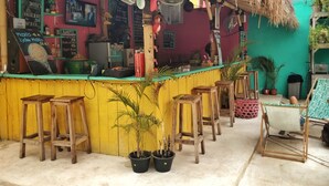 Un bar al aire libre colorido con taburetes de madera, techo de paja y varios letreros que anuncian bebidas.