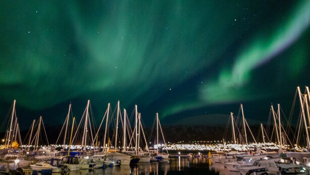 Een jachthaven in de nacht met aangemeerde boten en het Noorderlicht aan de hemel.