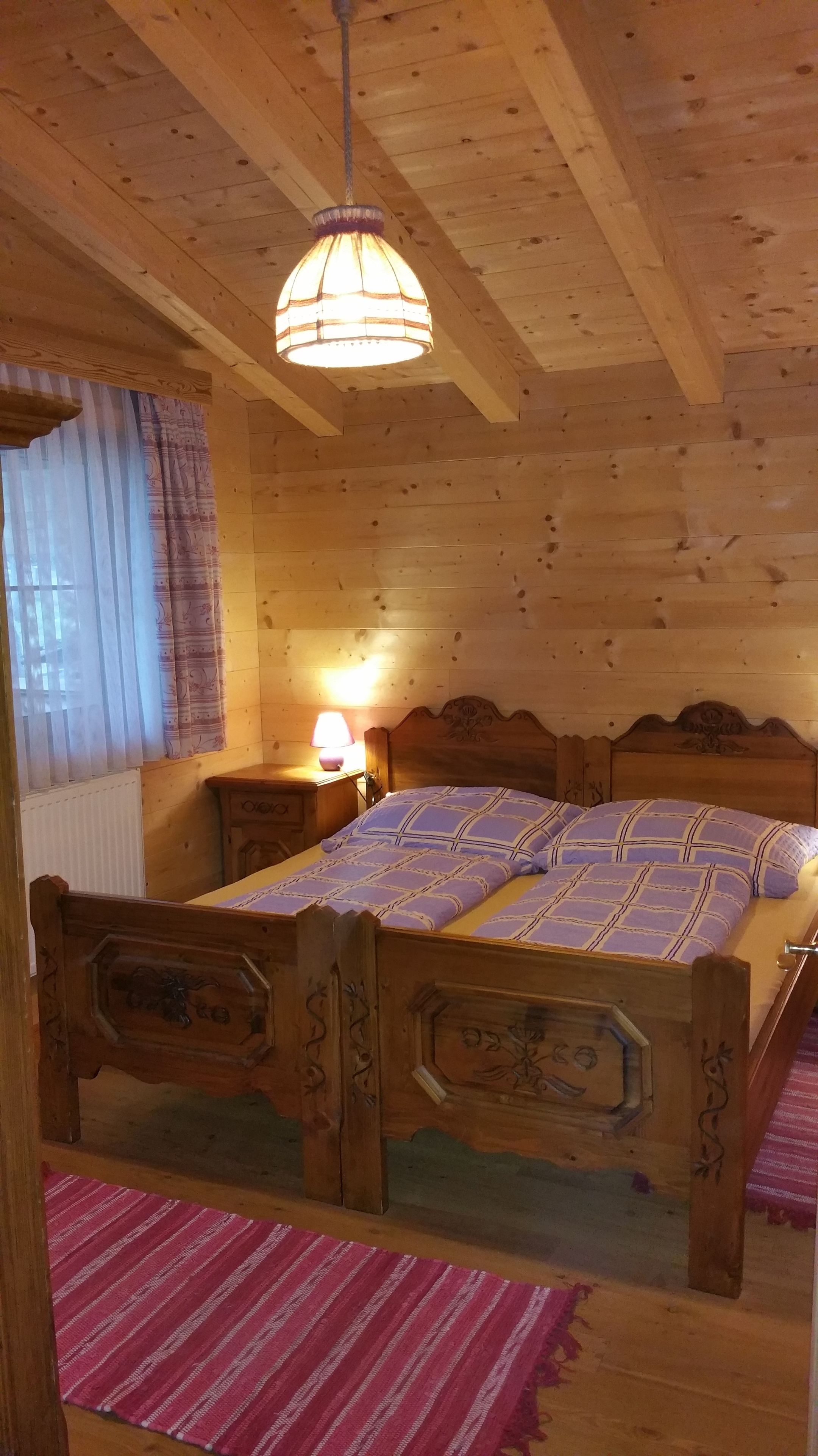 A wooden cabin room with a bed, a nightstand, a lamp, and a window with curtains.