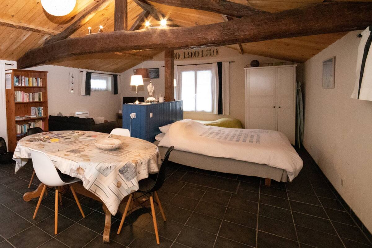 Une pièce chaleureuse avec un plafond en bois, un lit, une petite table recouverte d’une nappe ornée de motifs de journaux, une bibliothèque et une armoire blanche.