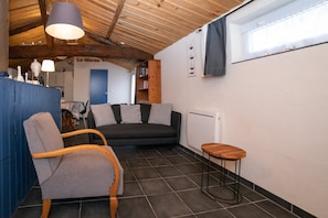 A cozy living room with a dark sofa, a wooden chair, a small wooden stool, and a blue cabinet. A large window with a curtain is on the right wall.