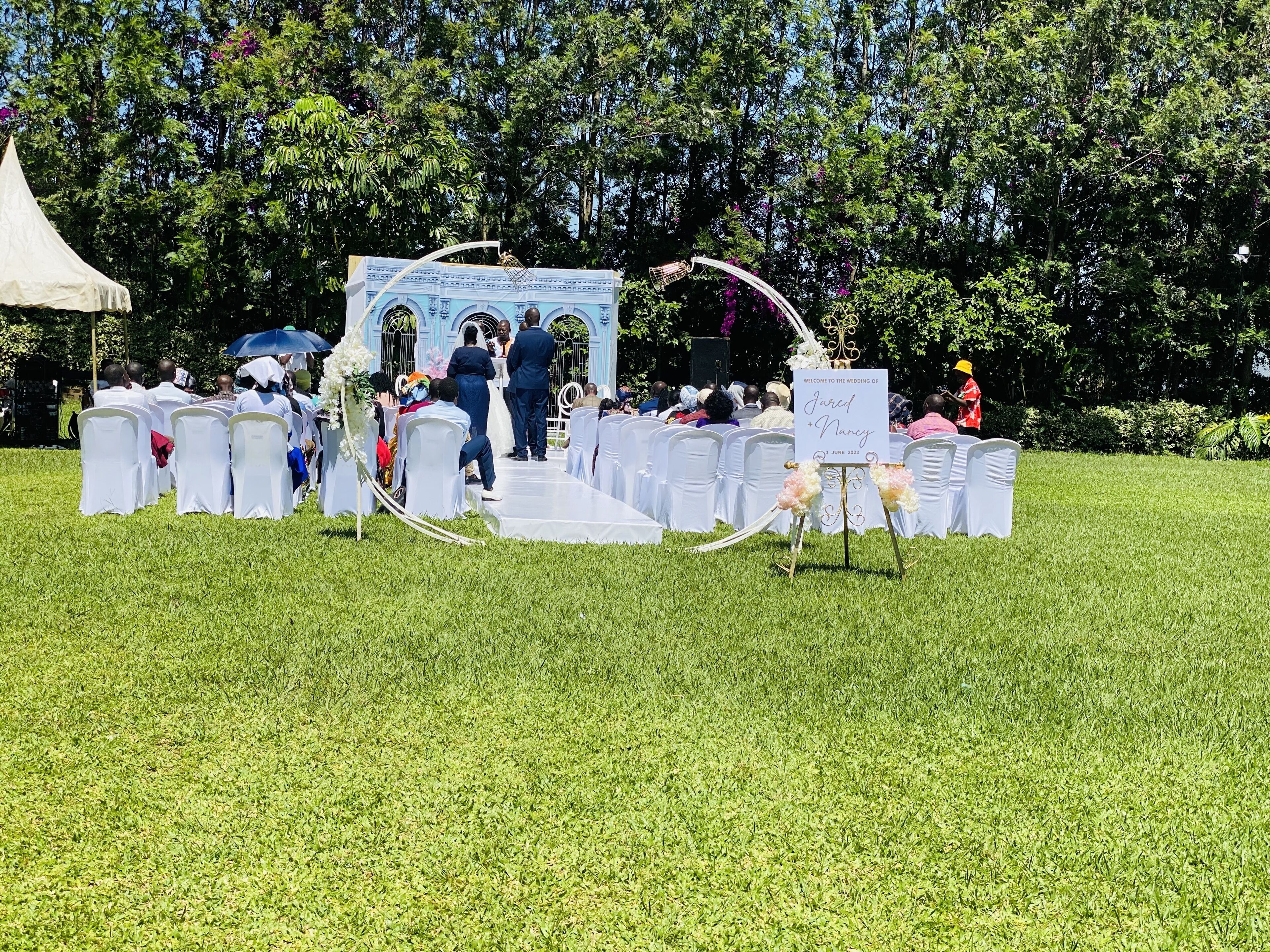 Eine Hochzeitszeremonie mit sitzenden Gästen auf weißen Stühlen, die in Reihen angeordnet sind, einer Bühne mit einer Transparente und einem Hintergrund aus Bäumen und Grünpflanzen.