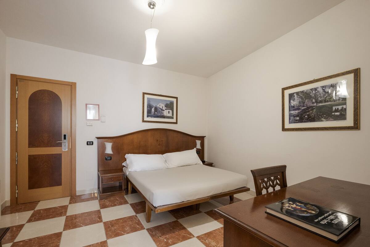 A hotel room with a wooden bed, a checkered floor, a wooden door, a nightstand, a table, and two framed pictures on the wall.