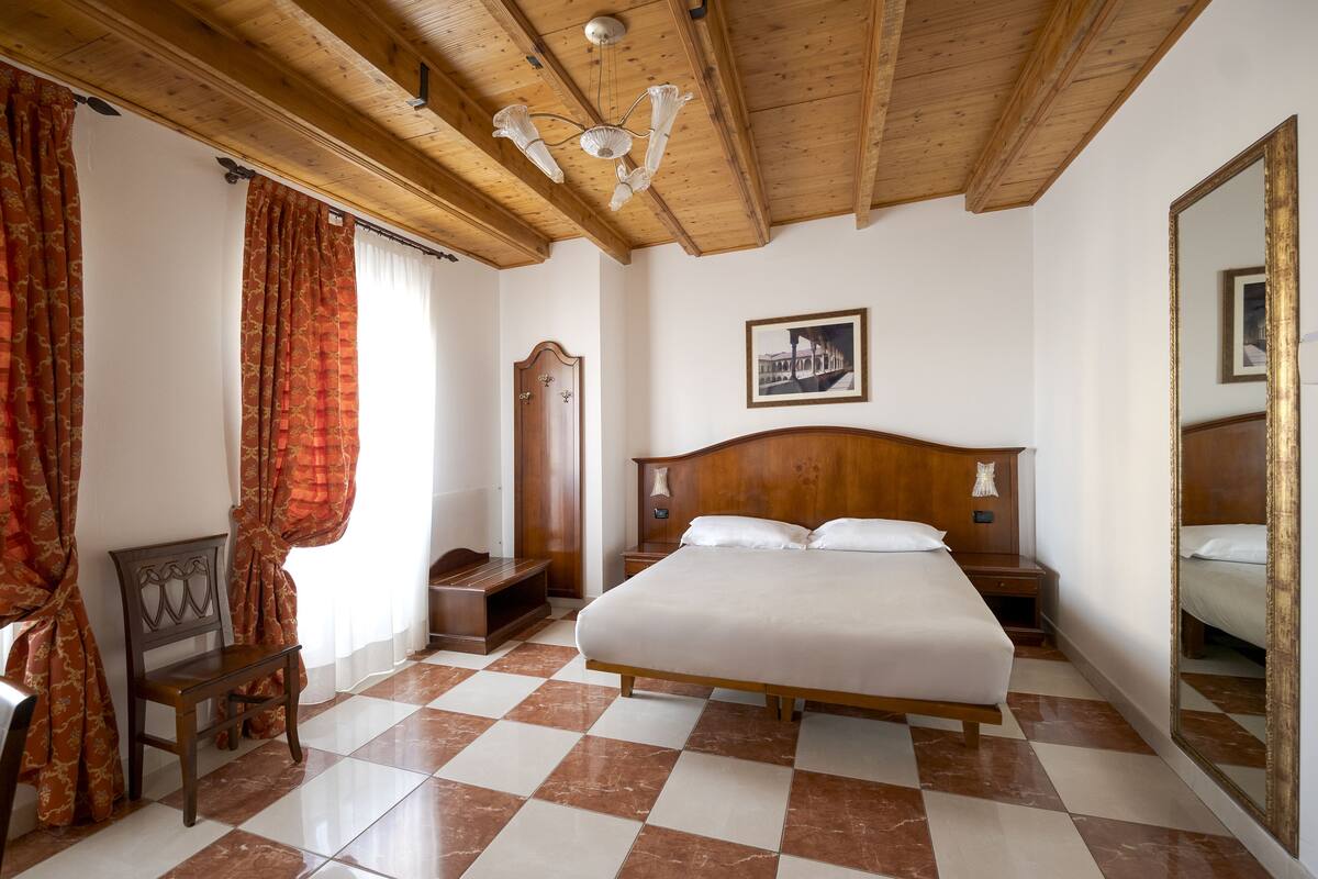 A bedroom with a large bed, wooden headboard, a nightstand, a bedside table, a chair, a wardrobe, and a mirror.