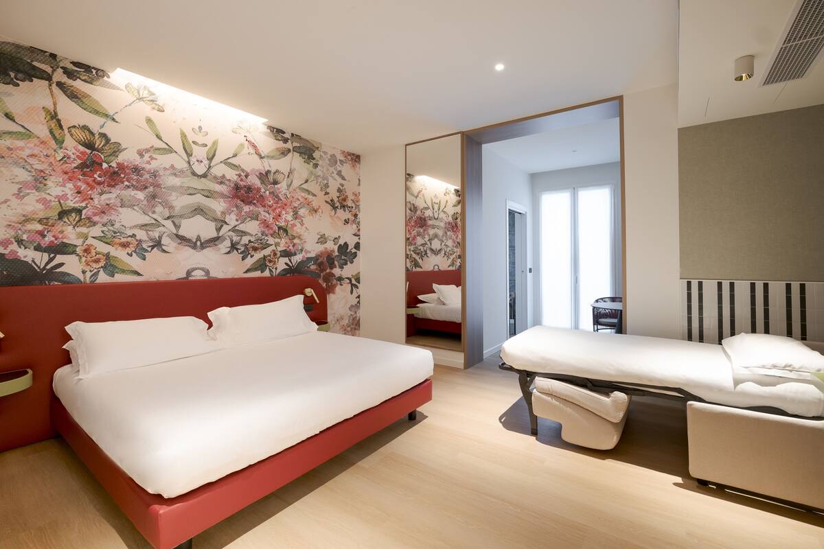 A modern bedroom with a large bed, a daybed, a mirror, and a floral wallpaper.