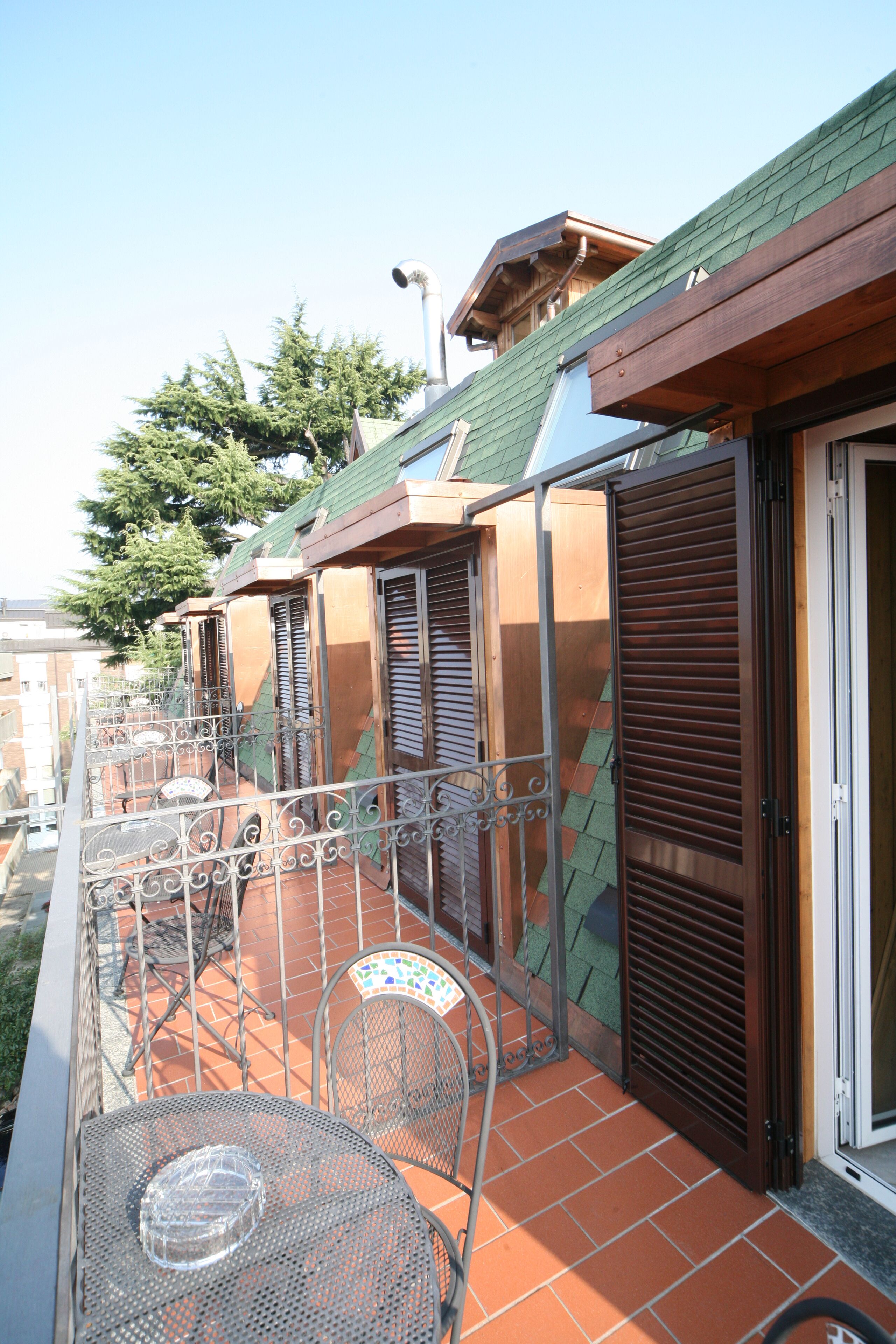 Un balcone con sedie e tavolo in metallo, pavimento piastrellato e un edificio con tetto verde e persiane in legno.