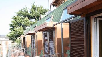 Un balcone con sedie e tavolo in metallo, pavimento piastrellato e un edificio con tetto verde e persiane in legno.