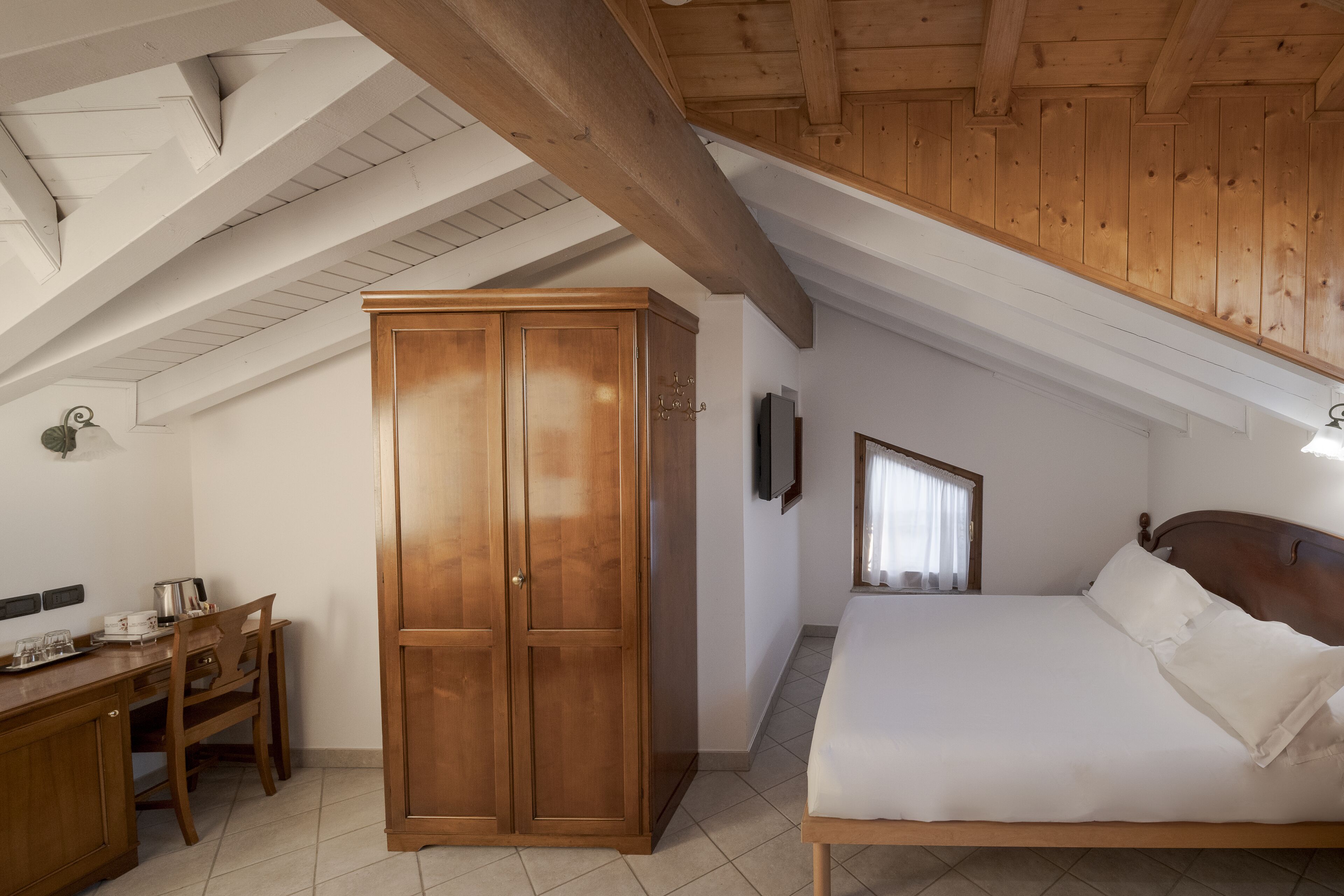 A bedroom with a wooden wardrobe, a bed with white bedding, a desk with a chair, and a television on the wall.