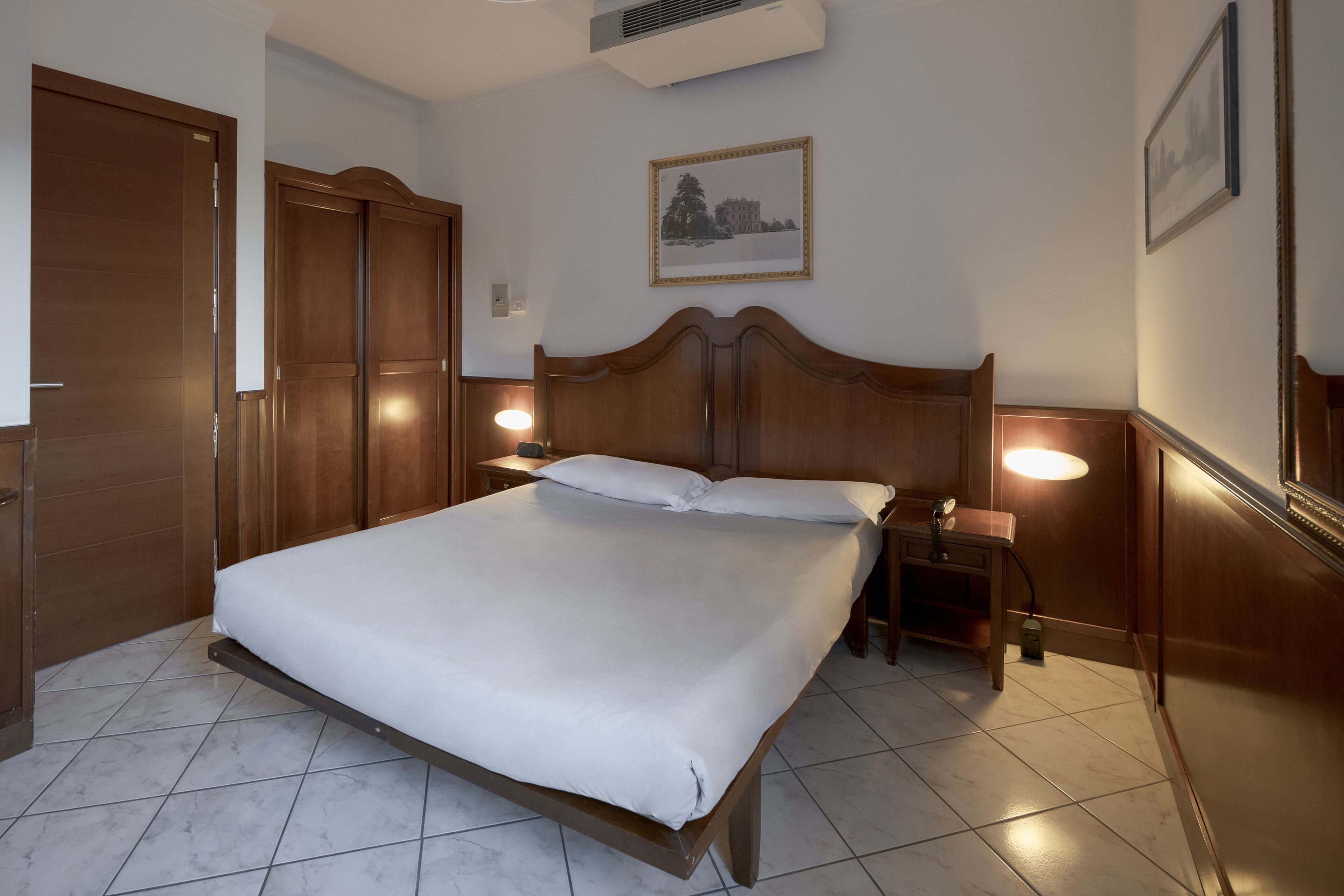 A neatly arranged bedroom with a large bed, wooden wardrobe, bedside lamps, and a framed picture on the wall.