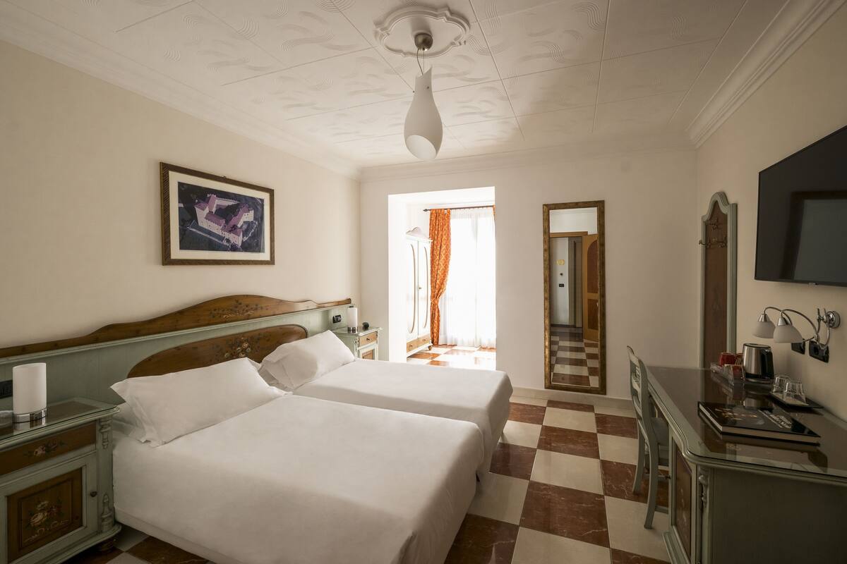 A hotel room with two beds, a television, a desk with a lamp, and a framed picture on the wall.