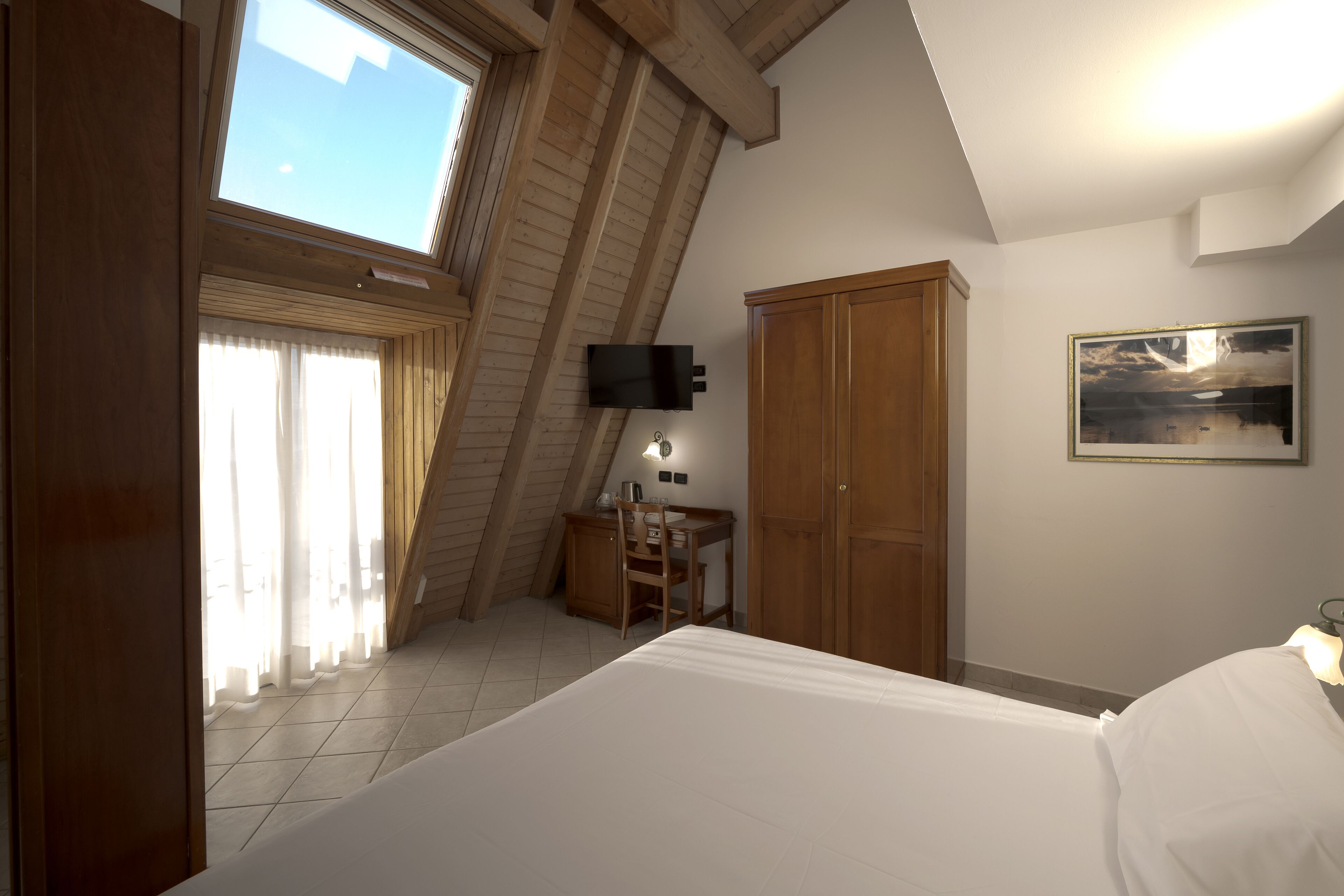 A bedroom with a sloped ceiling, a bed, a desk, a chair, a wardrobe, and a painting on the wall.