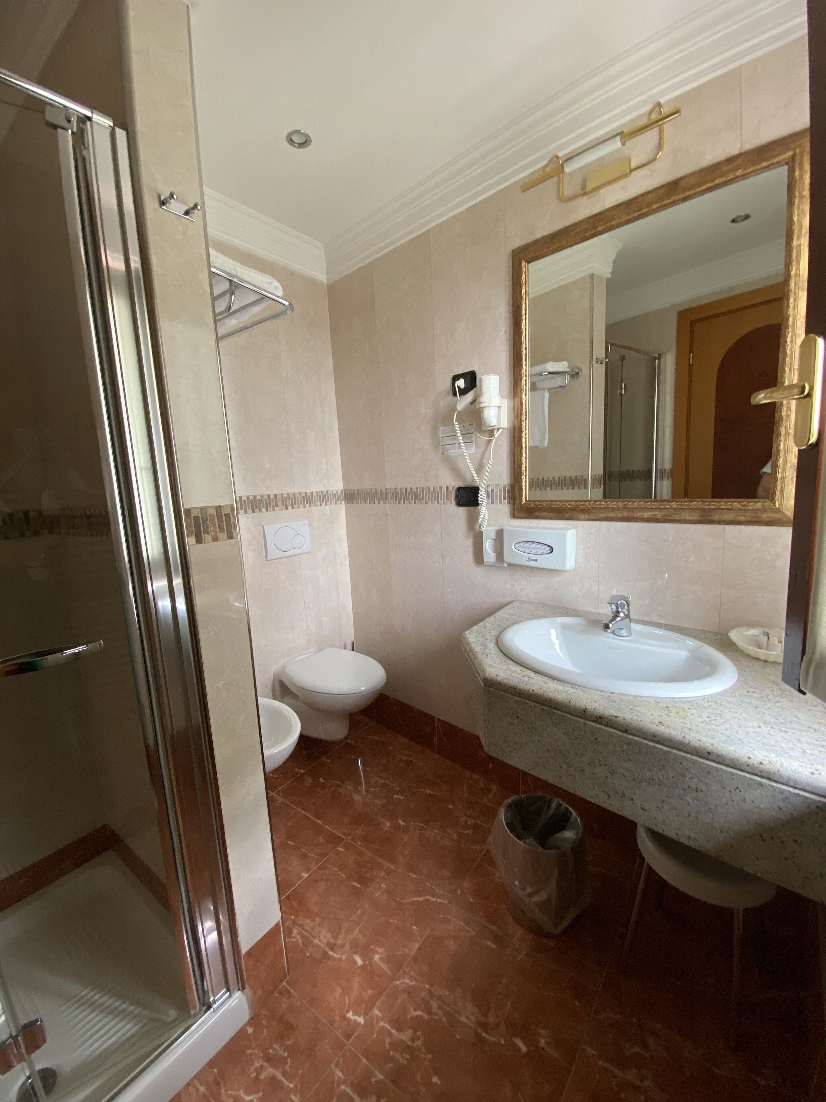 A bathroom with a glass shower enclosure, a white toilet, a sink with a mirror, and a trash bin.