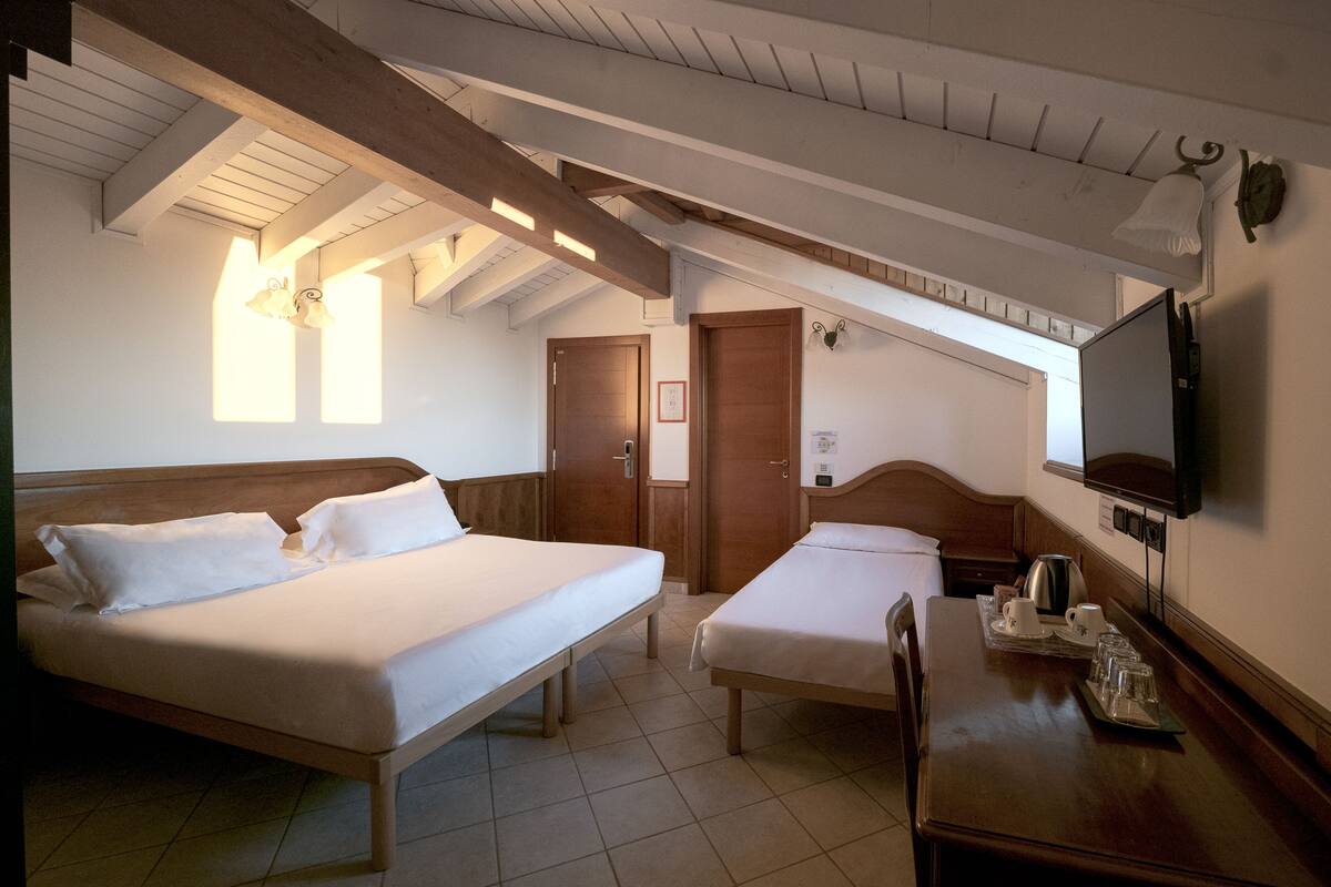 A hotel room with two beds, a wooden table with a tray of glasses and cups, a television, and a ceiling with exposed beams.