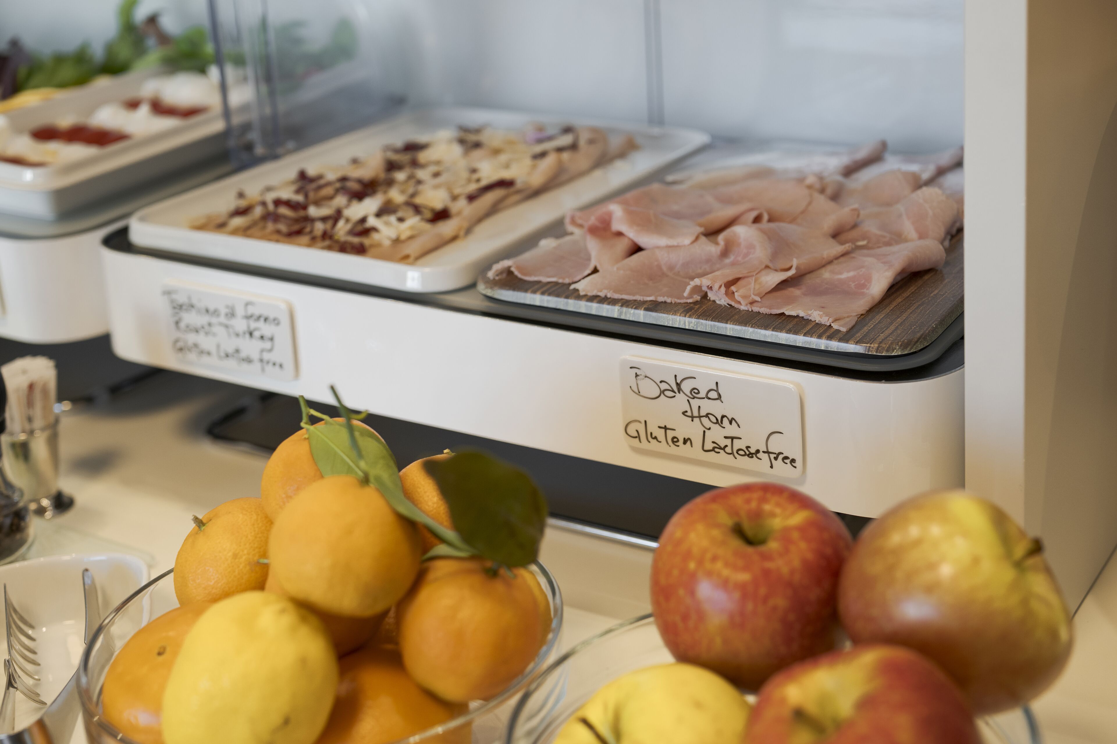A buffet with various food items including baked ham, gluten-free options, and fresh fruits like oranges and apples.