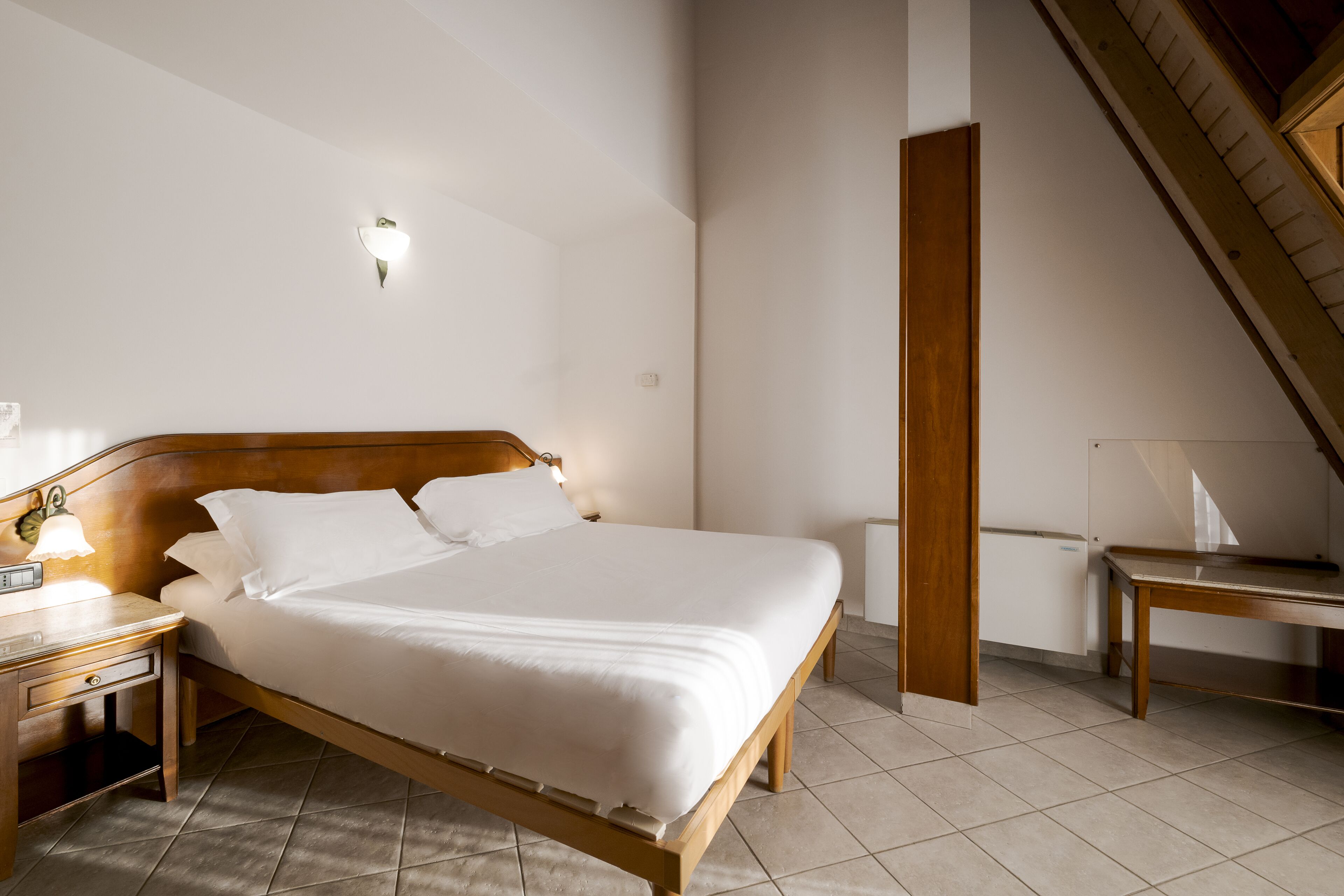 Ein ordentlich bezogenes Bett in einem Hotelzimmer mit einem hölzernen Kopfteil, einem Nachttisch mit Lampe und einem kleinen Tisch mit Stuhl.