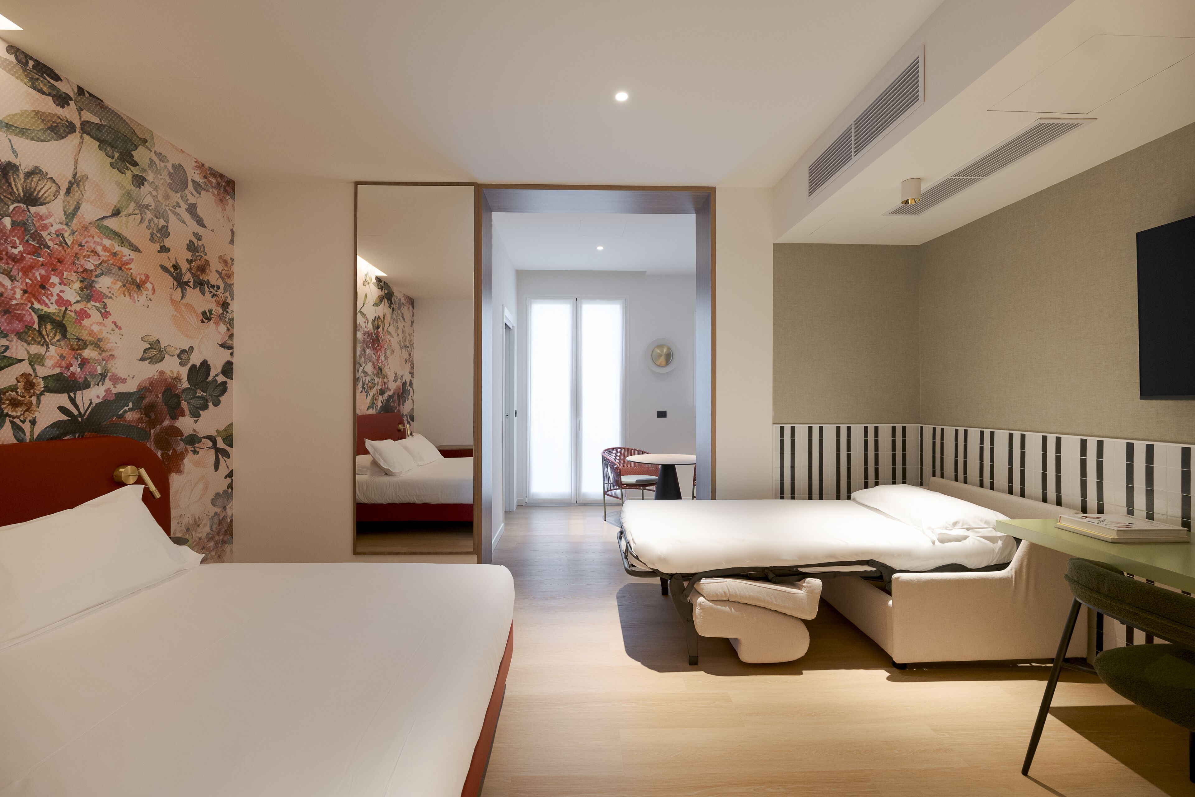 A hotel room with a bed, a sofa bed, a small table, and a TV. There is a door leading to another room and a window with blinds.