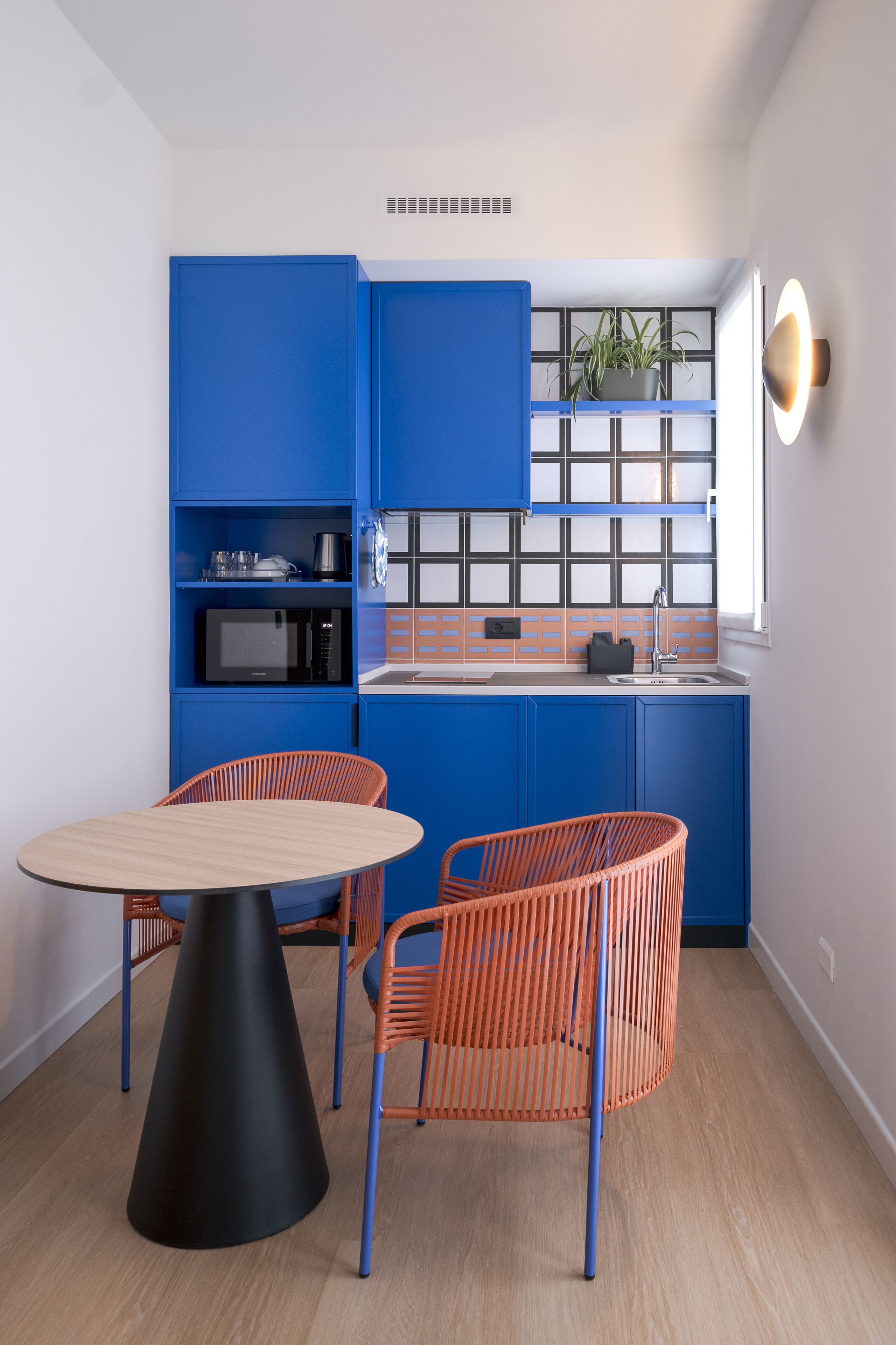 A compact kitchen with blue cabinets, a microwave, and a sink. There is a round table with a black cone base and two orange chairs.