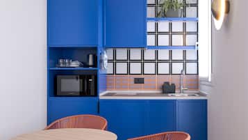 A compact kitchen with blue cabinets, a microwave, and a sink. There is a round table with a black cone base and two orange chairs.