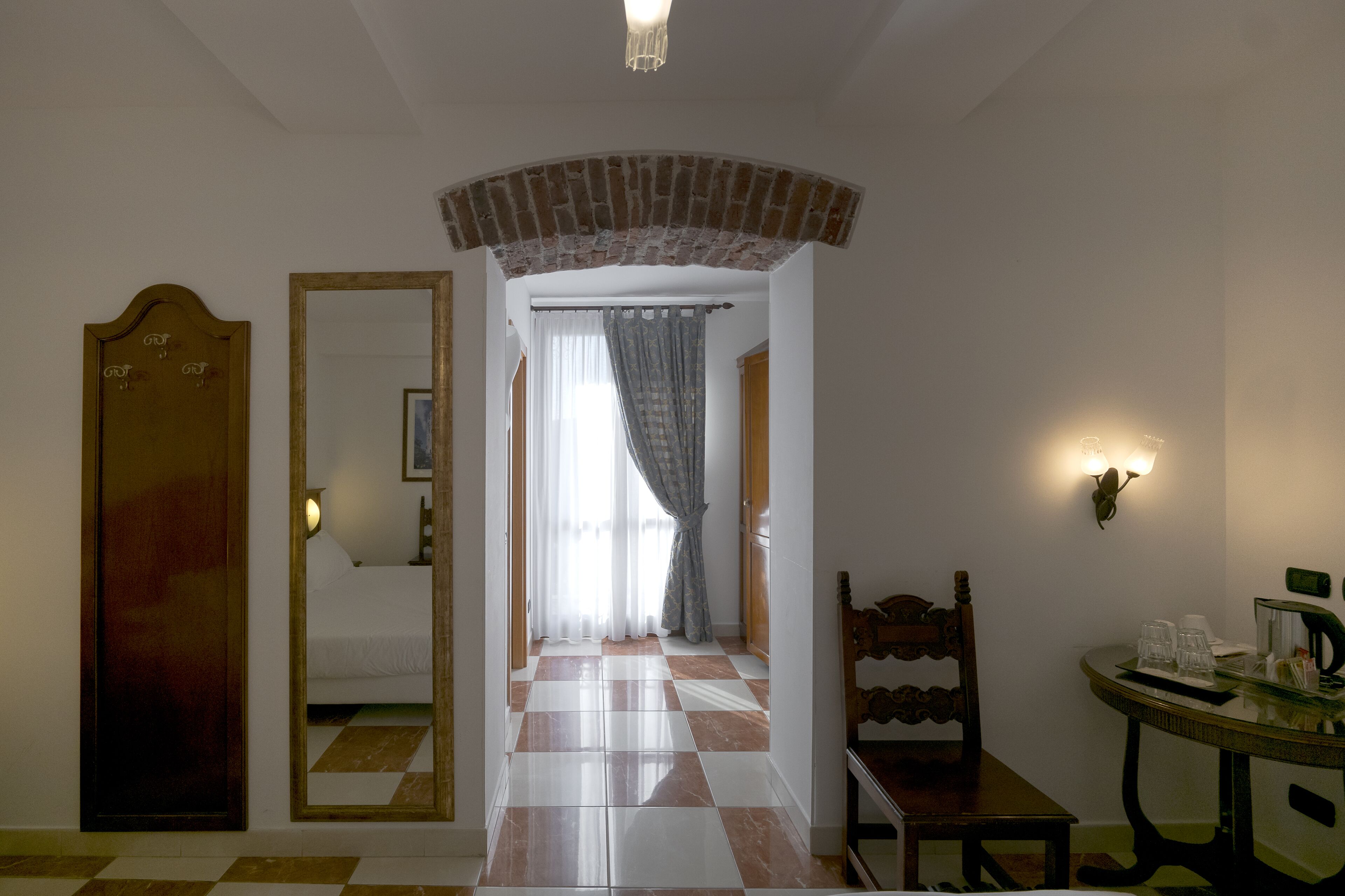 A hotel room with a checkered floor, a wooden door, a mirror, a bed, a table with a chair, and a wall-mounted light fixture.