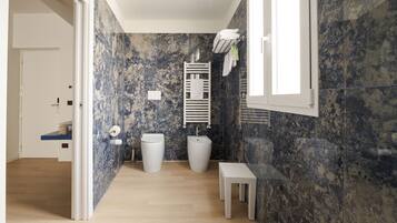 A bathroom with a marble wall, a toilet, a bidet, a towel rack, and a small table.
