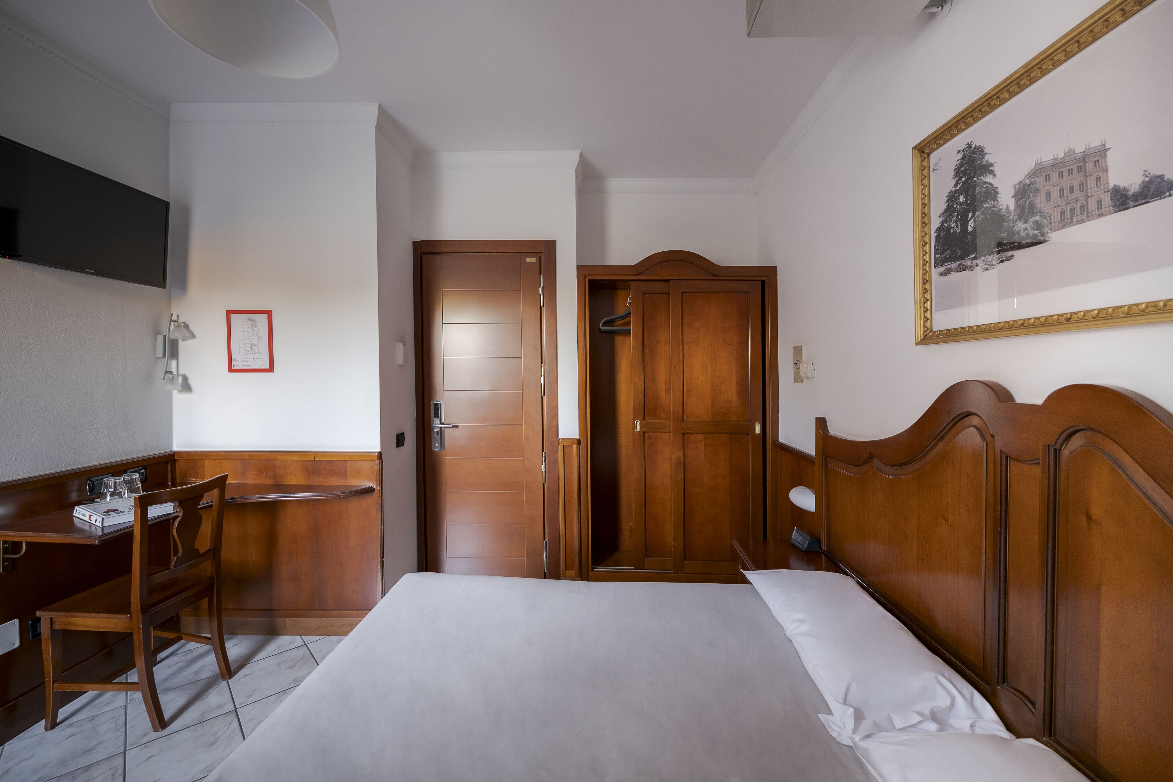 Habitación de hotel con una cama, un escritorio con una silla, un armario, un cuadro en la pared y un televisor montado en la pared.
