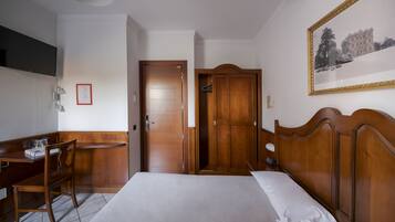 Habitación de hotel con una cama, un escritorio con una silla, un armario, un cuadro en la pared y un televisor montado en la pared.