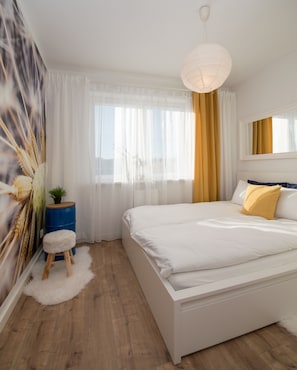 A modern bedroom with a large bed, a bedside table with a plant, a fluffy rug, and a view of the outdoors through sheer curtains. - Fresh Apart Marina (Bydgoszcz)