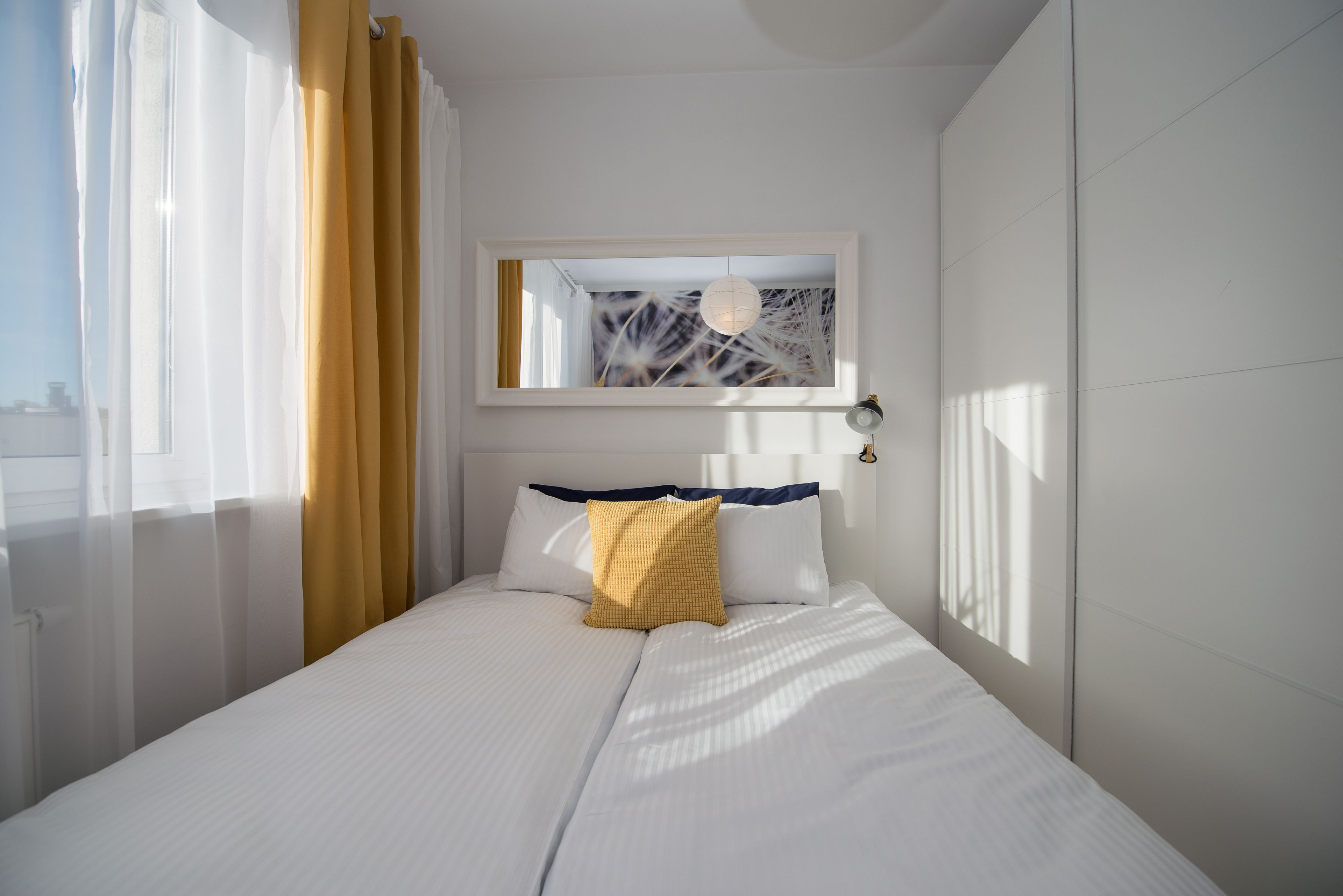 Un lit bien fait, avec du linge de lit blanc et un seul oreiller jaune, une table de chevet avec une lampe, et une fenêtre ornée de rideaux légers et d’un voilage jaune.