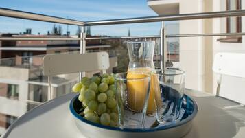 A table with a glass pitcher, a glass of orange juice, and a bowl of green grapes on a balcony with a clear view of buildings in the background.