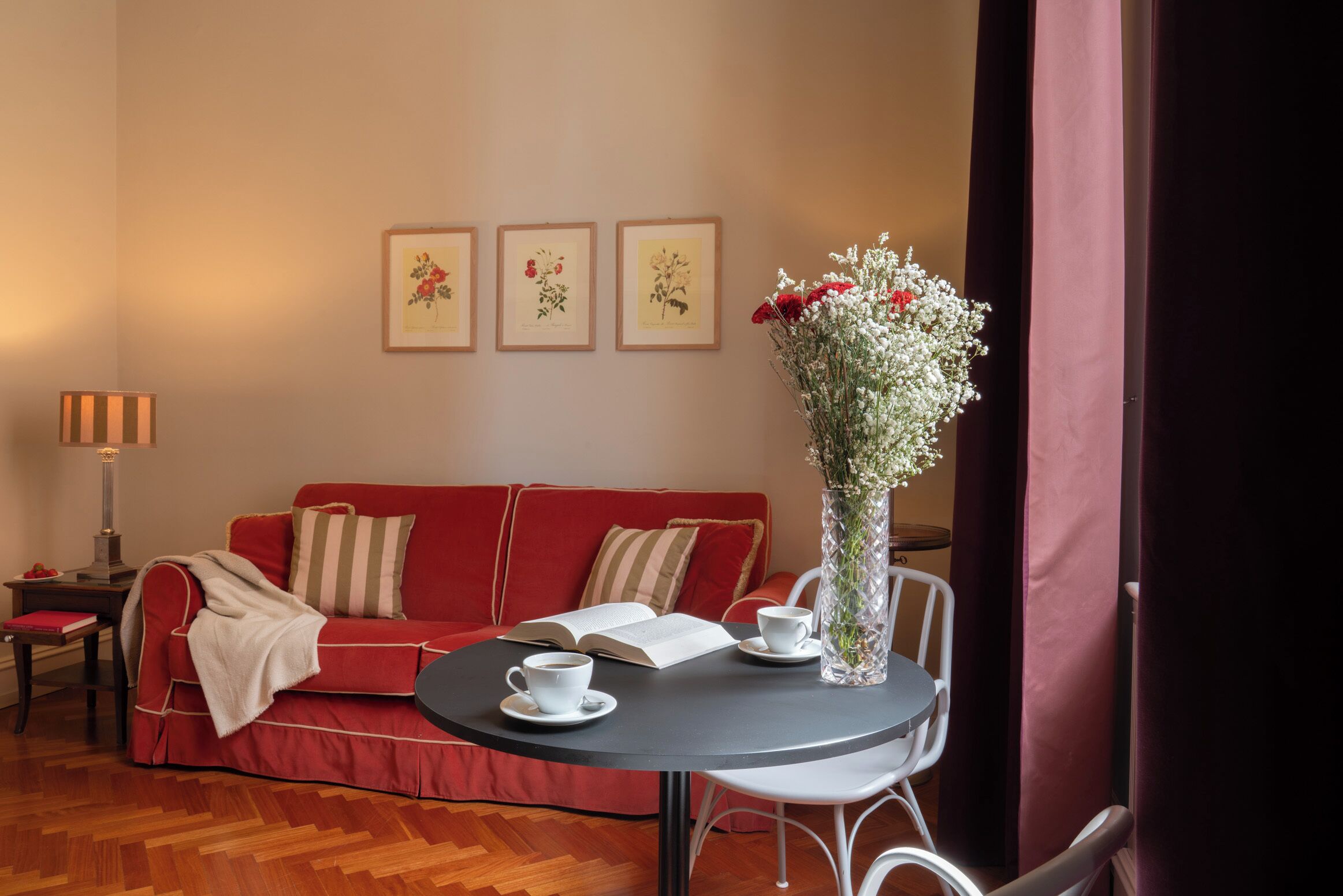 Una habitación acogedora con un sofá rojo, una mesa redonda con un florero y grabados botánicos enmarcados en la pared.