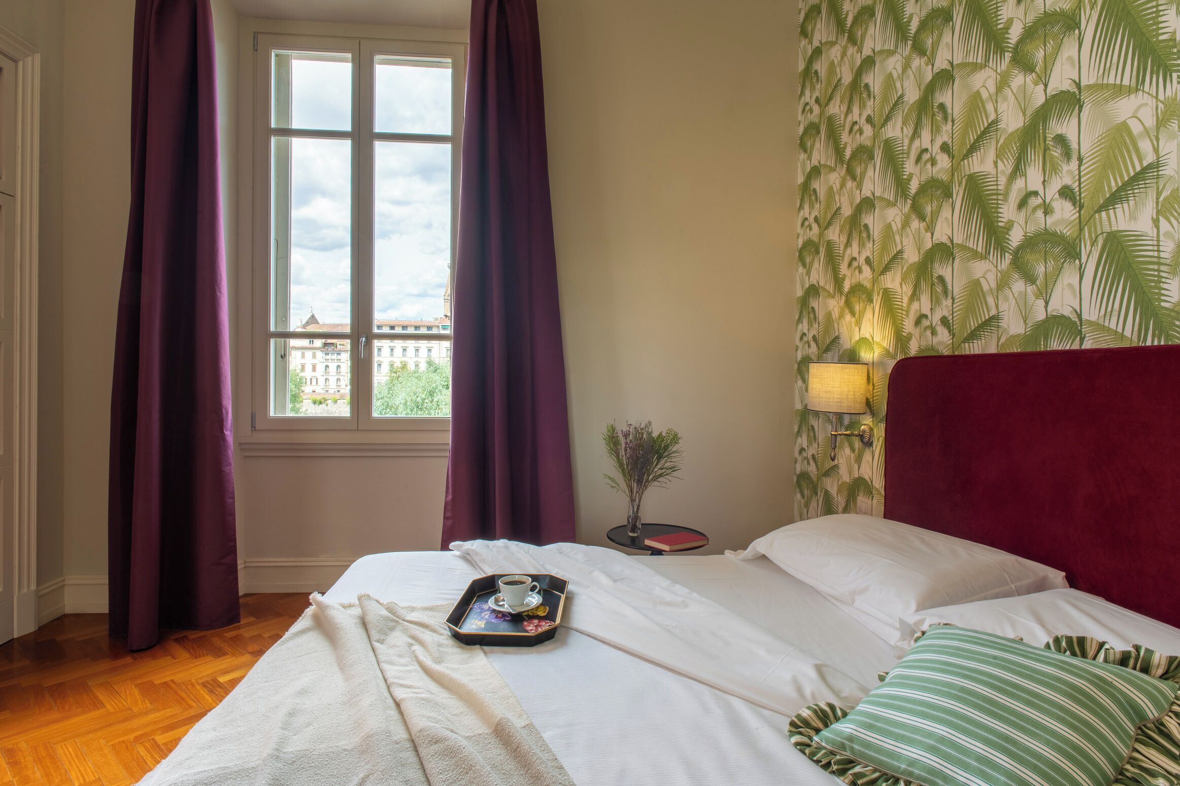 Une chambre d’hôtel avec un lit, une fenêtre avec des rideaux, une table de chevet avec une lampe et une vue sur les bâtiments à l’extérieur.