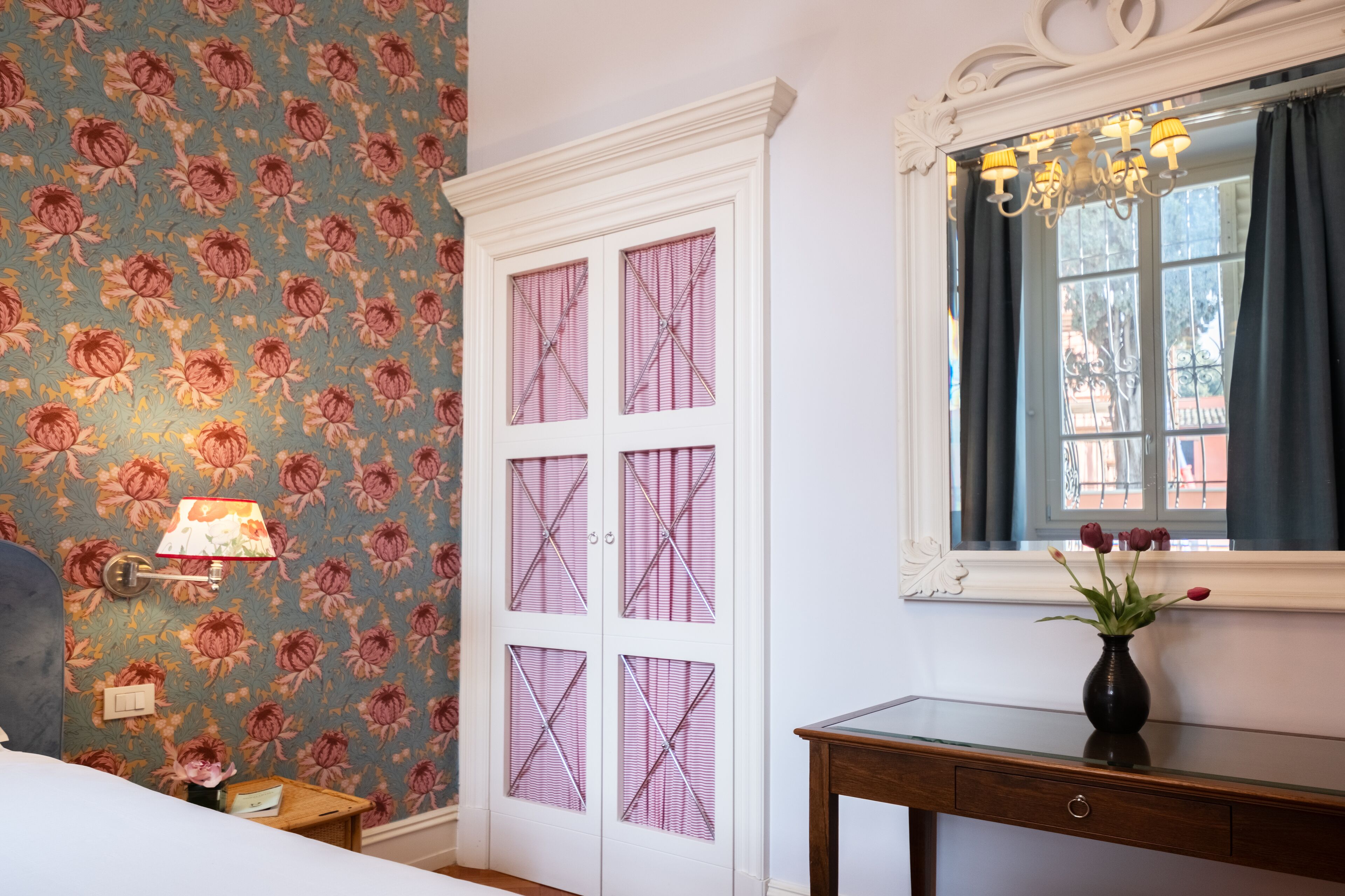A room with floral wallpaper, a white door with glass panels, a bedside table with a lamp, and a window with a view.