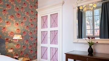 A room with floral wallpaper, a white door with glass panels, a bedside table with a lamp, and a window with a view.