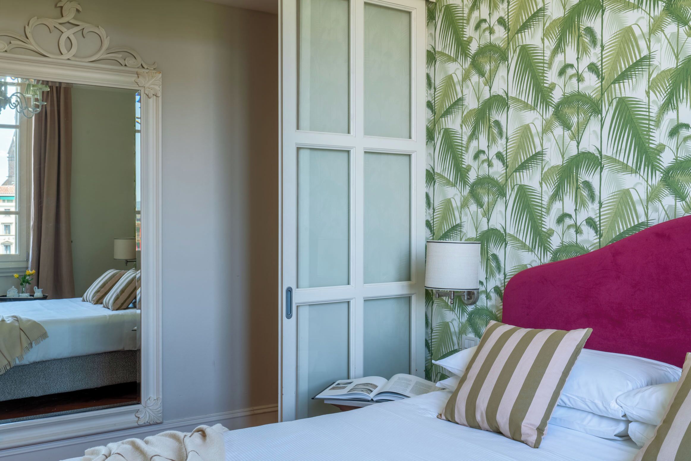 A hotel room with a bed, a mirror, a nightstand with a lamp, and a window with curtains.