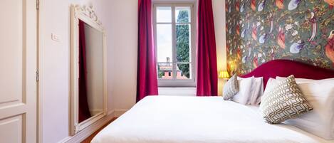 A hotel room with a large bed, a window with curtains, a mirror, and a floral wallpaper.