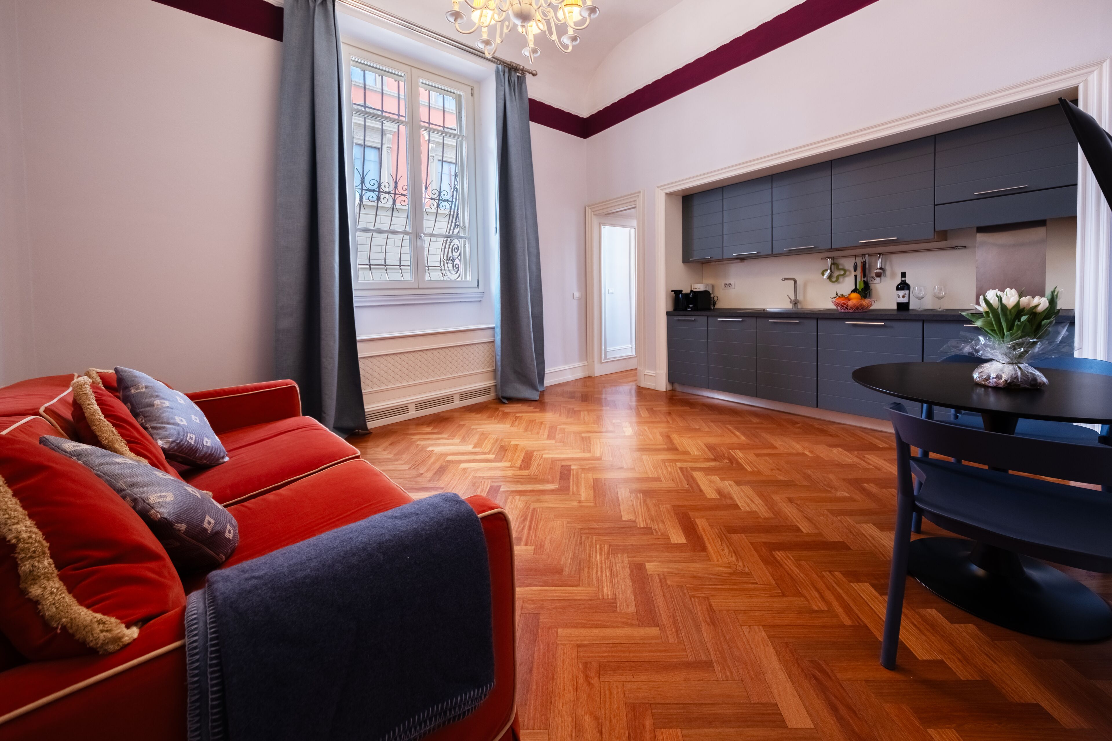 A modern living room with a red sofa, wooden parquet flooring, and a dining area with a table and chairs.