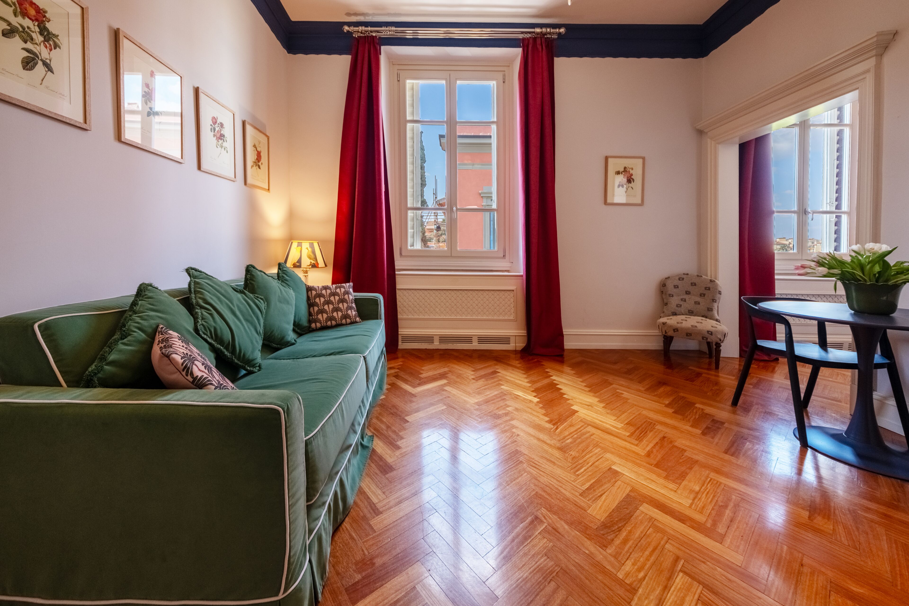 A room with a green sofa, a small table with a plant, a chair, a window with red curtains, and a radiator.
