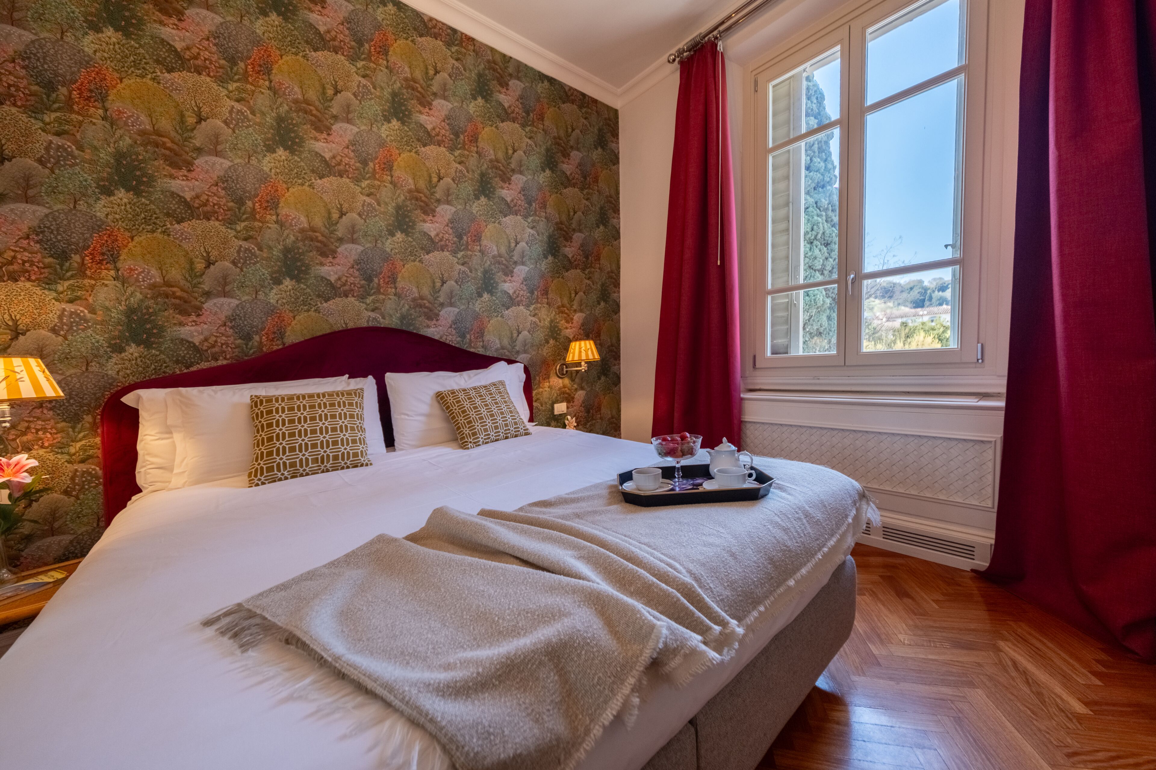 Un letto rifatto con cura, un vassoio con tè e tazze, una tenda rossa e una finestra vista alberi.