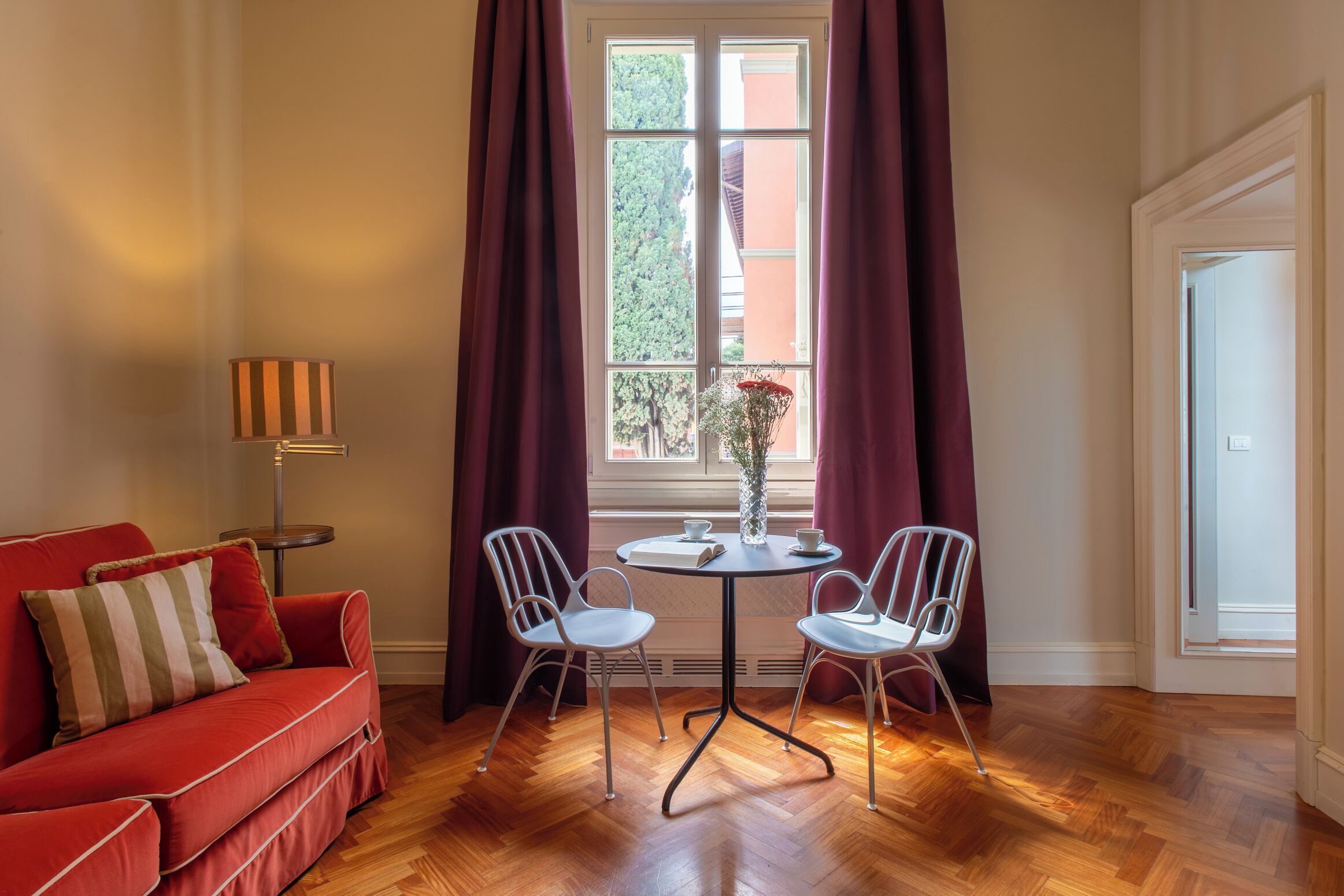 A room with a red sofa, two white chairs, a small round table, a floor lamp, and a window with curtains.