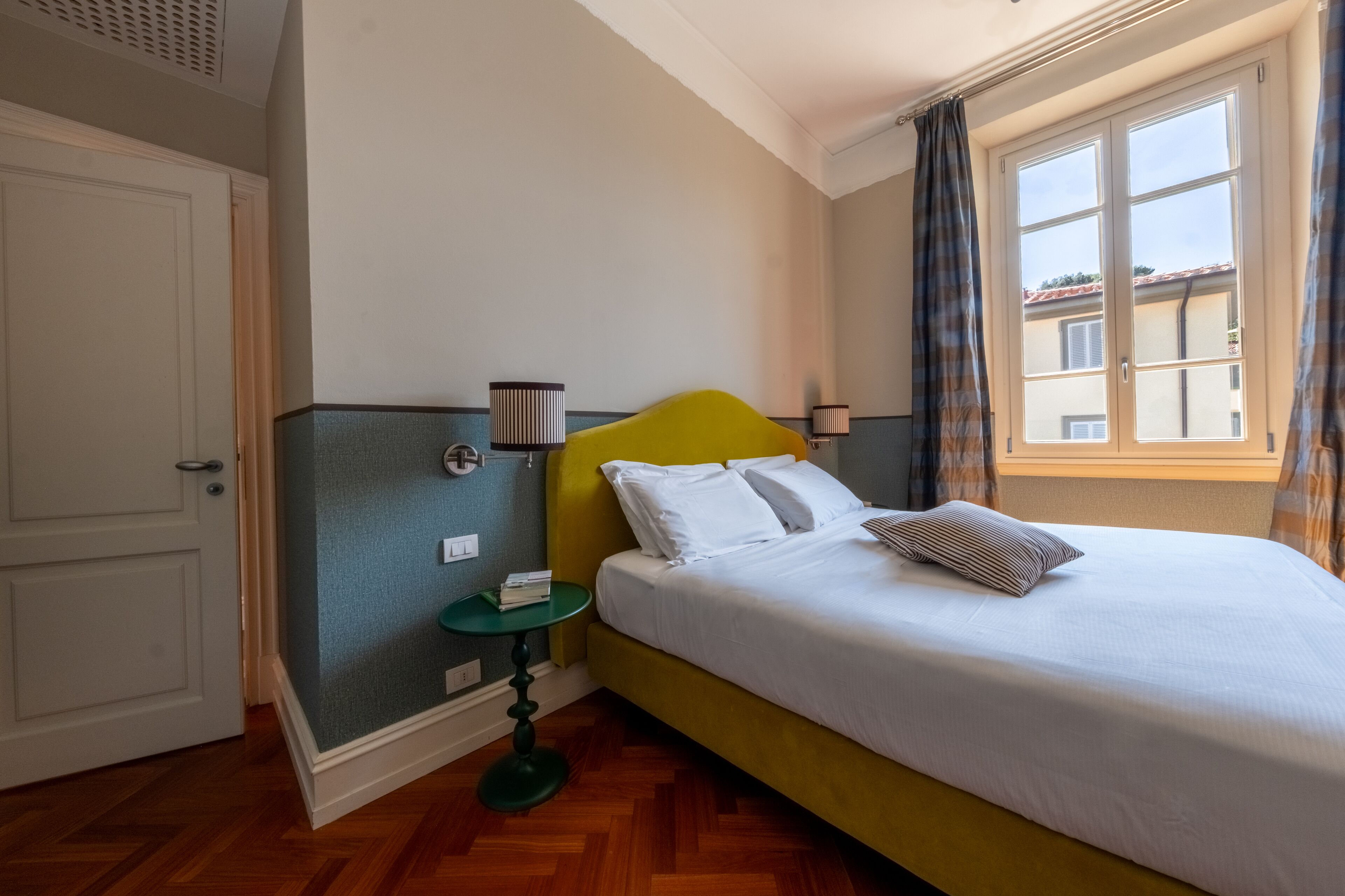 A hotel room with a bed, a nightstand, a window with curtains, and a door.