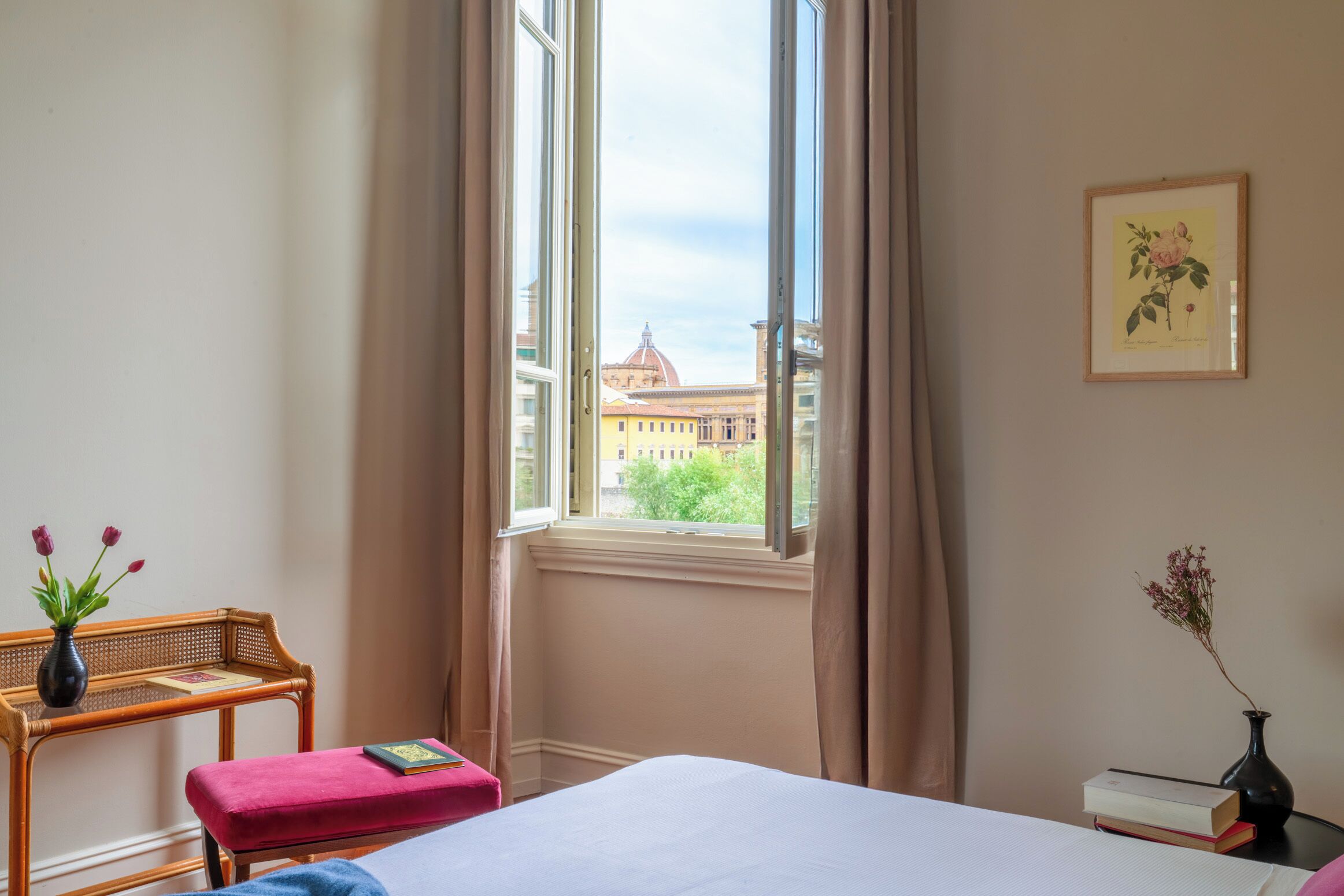 A room with a bed, a window with curtains, a small table with a vase, and a framed picture on the wall.