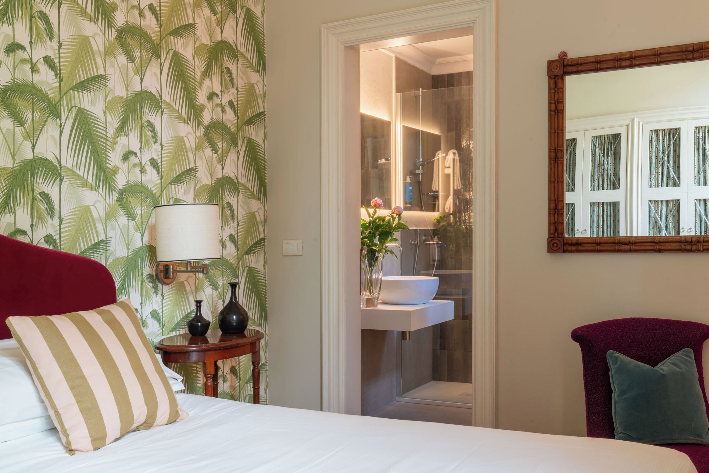 Une chambre d’hôtel avec un lit, une table de chevet, une lampe, un miroir et une salle de bain visible à travers une porte ouverte.