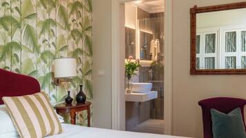 Une chambre d’hôtel avec un lit, une table de chevet, une lampe, un miroir et une salle de bain visible à travers une porte ouverte.