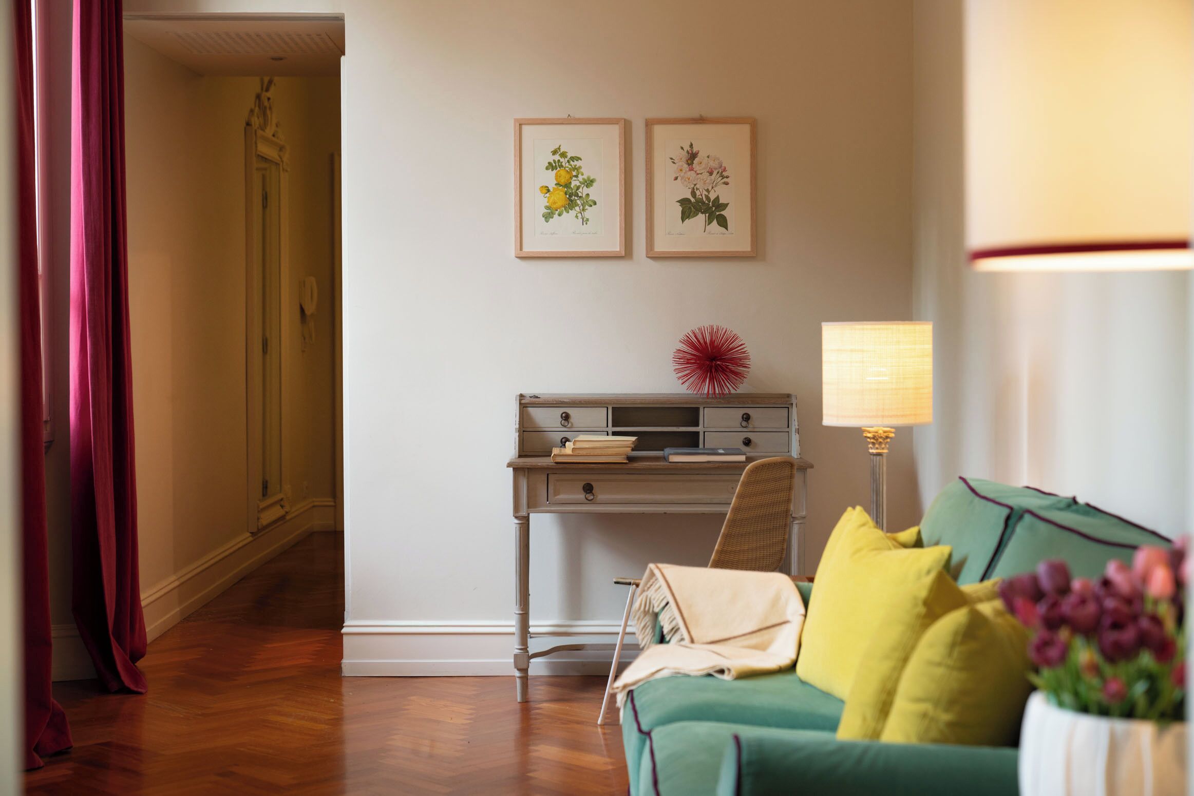 Ein Zimmer mit Schreibtisch, Stuhl, Lampe und einer Couch. An der Wand befinden sich zwei gerahmte Pflanzenbilder.