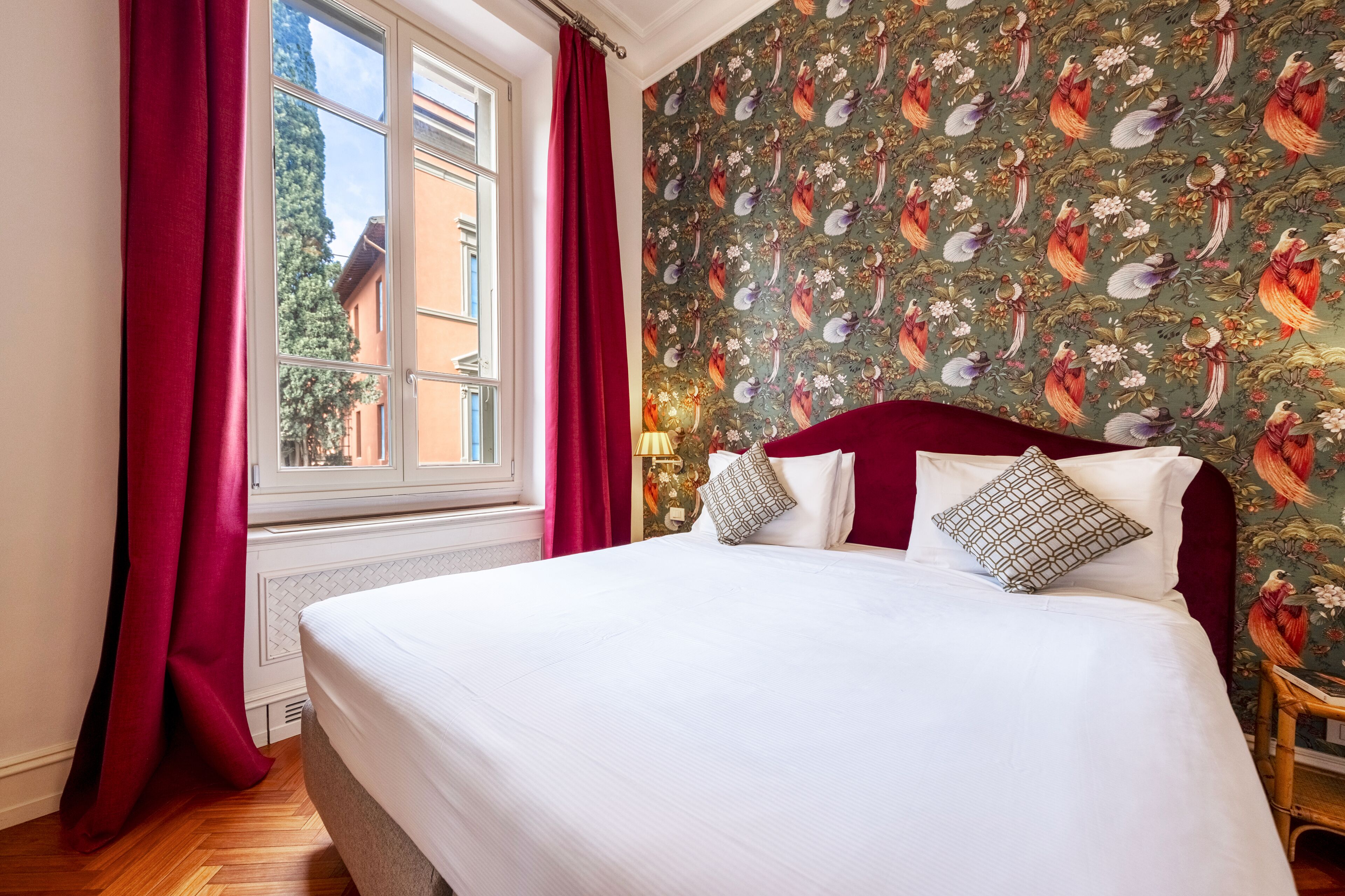 A hotel room with a large bed, red curtains, a floral wallpaper, and a view of buildings outside the window.
