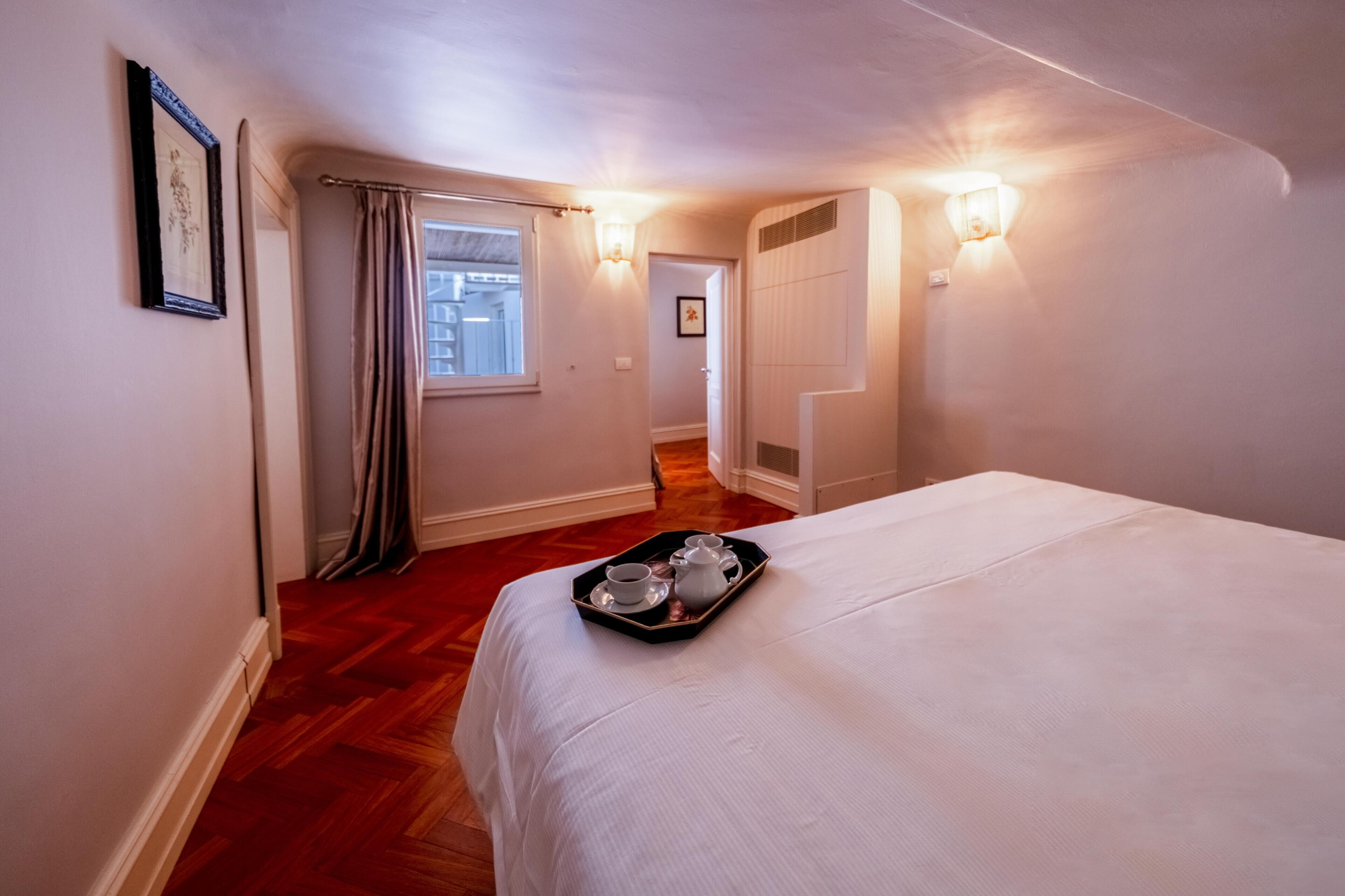 Ein Hotelzimmer mit einem Bett, einem Tablett mit Tee und Tassen, einem Fenster mit Vorhängen und Wandleuchten.