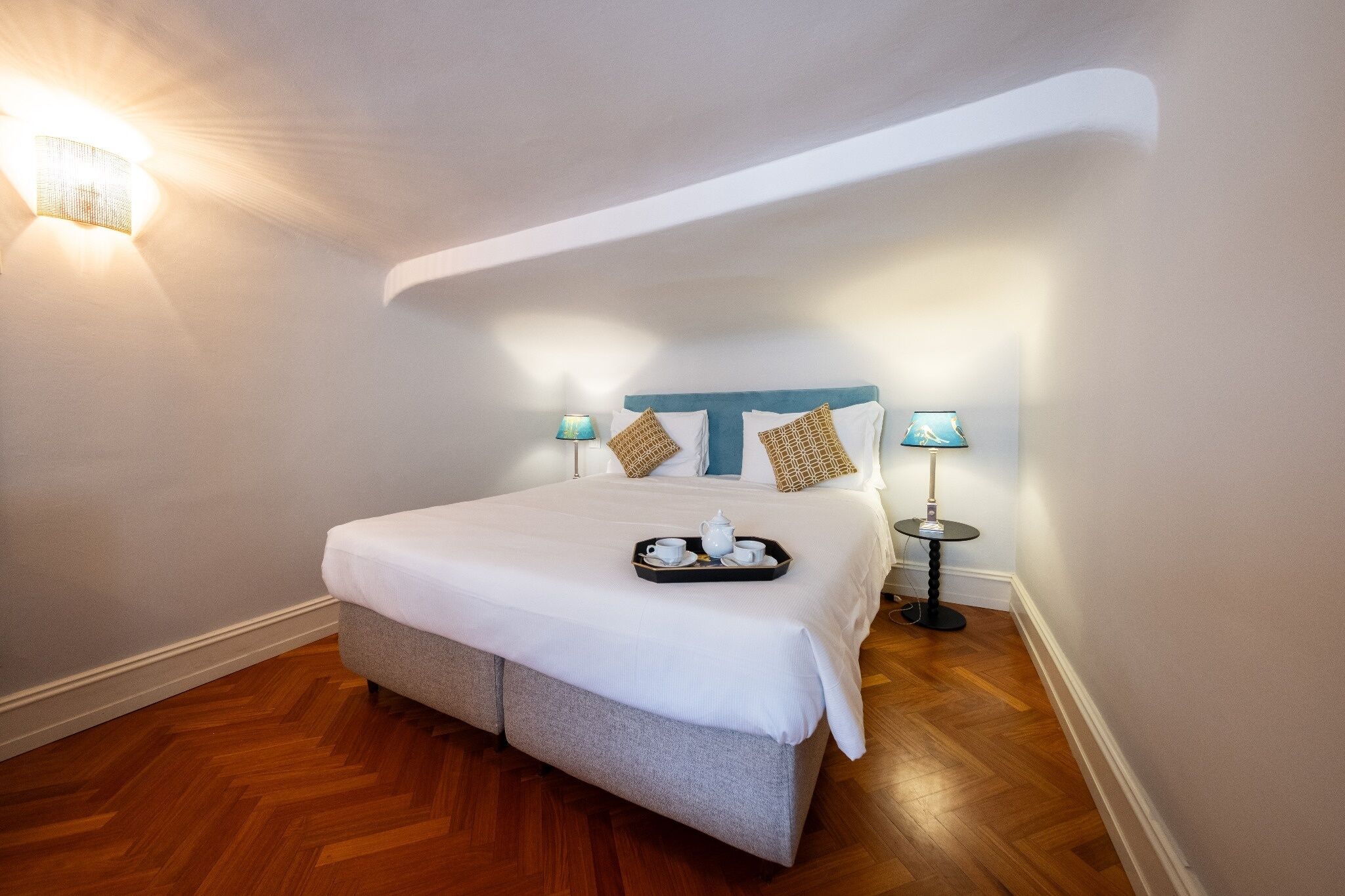 A neatly made bed with a tray of tea and cups, two bedside lamps, and a wooden floor.