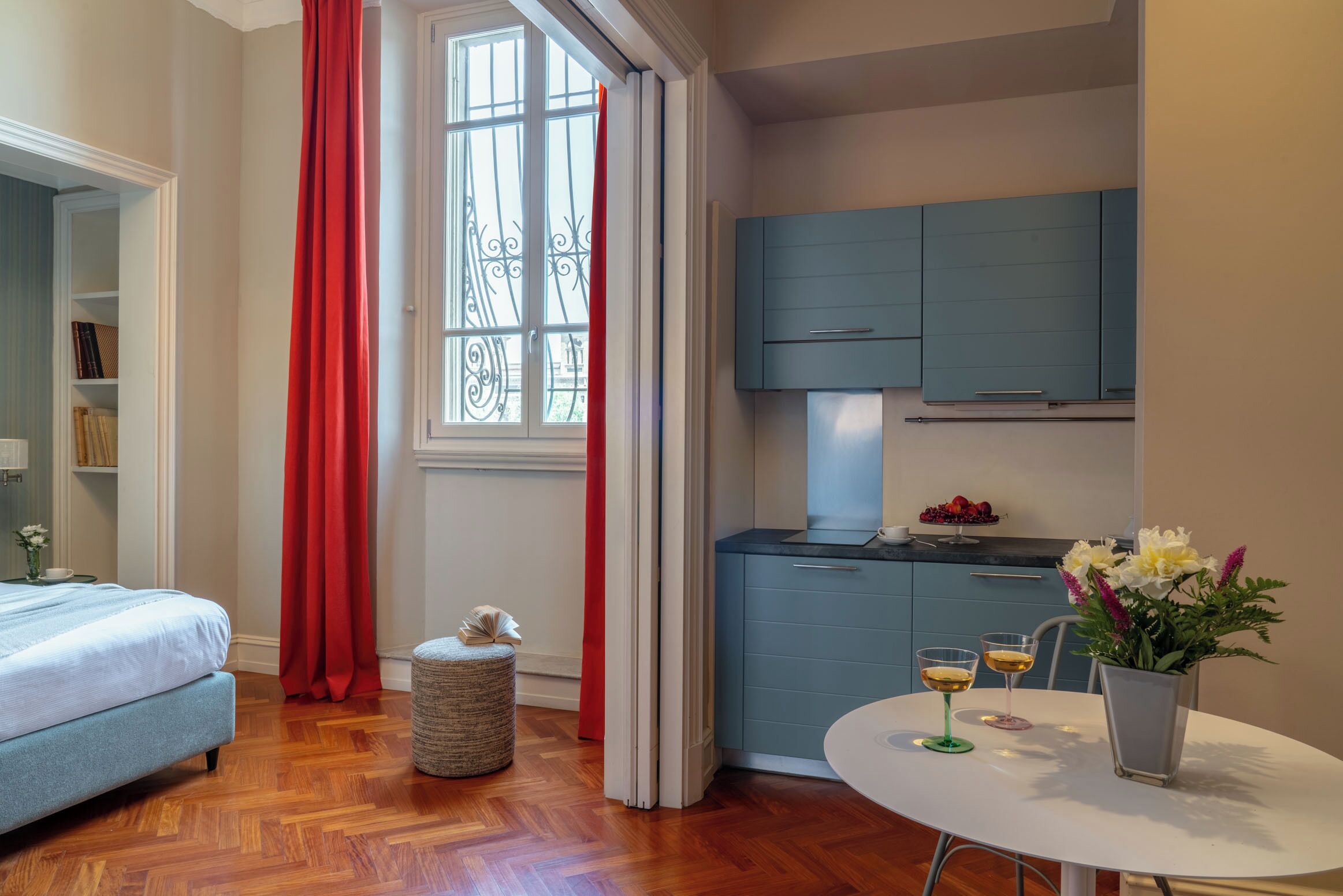 Une chambre à coucher de taille modeste, comprenant un lit, un petit pouf rond, une table à manger avec deux verres et un vase de fleurs.