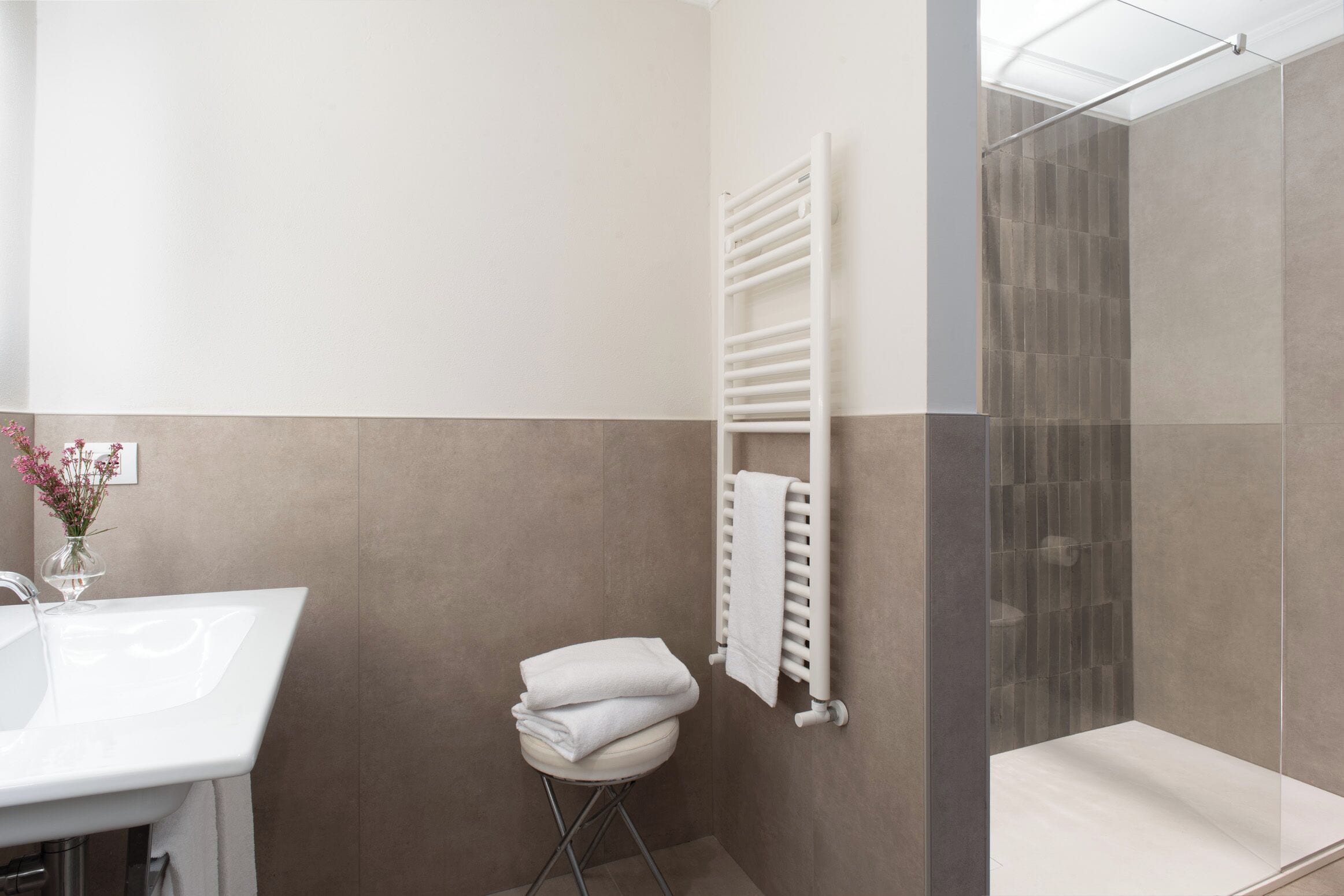 A bathroom with a white sink, a towel rack, a glass shower enclosure, and a small stool.